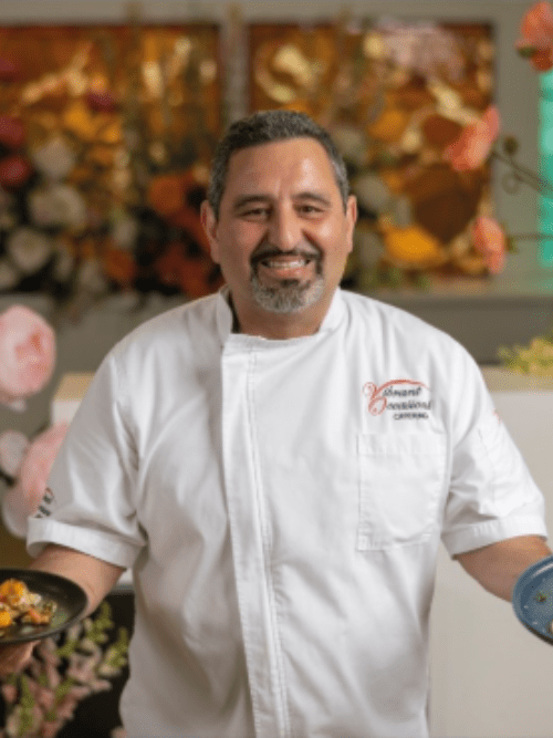 Chef Serge Krikorian A smiling chef in a white uniform holds plates of food in both hands, standing in front of a colorful background with flowers.