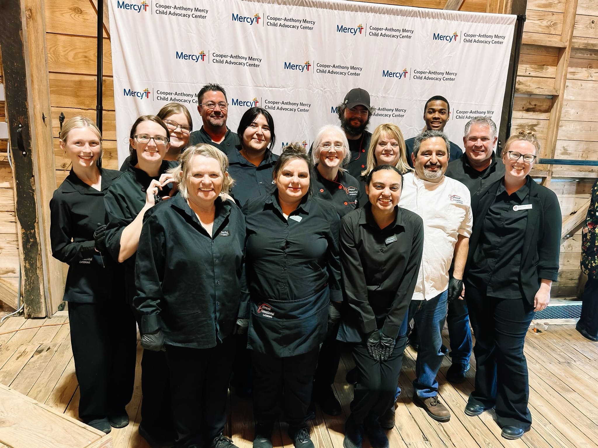 A group of thirteen smiling people in black and white uniforms pose together indoors on a wooden floor, in front of a step-and-repeat banner featuring Mercy and a Child Advocacy Center logo.