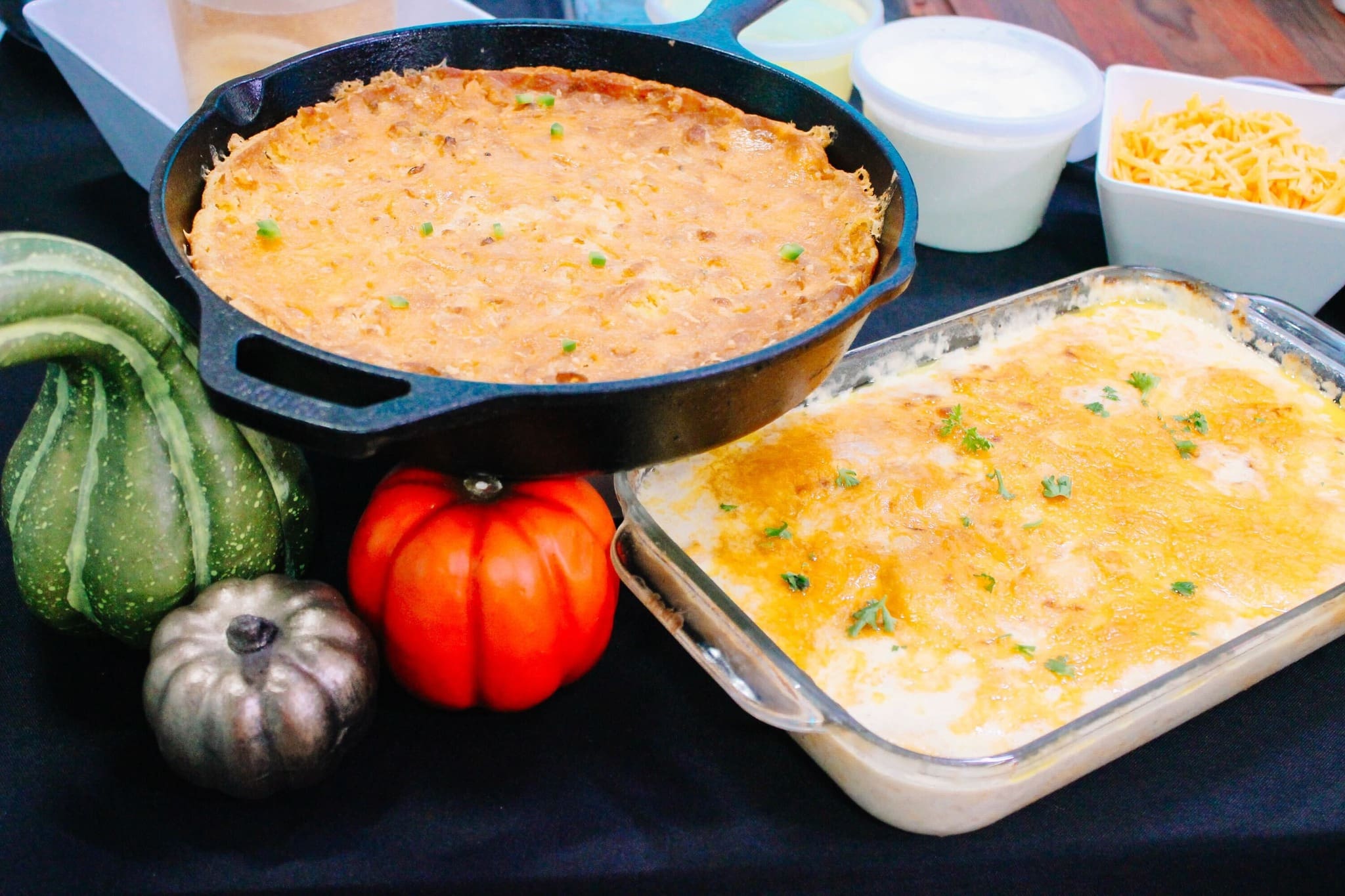 A skillet and a glass baking dish filled with baked casseroles sit on a table, surrounded by three decorative gourds, a bowl of shredded cheese, and a bowl of sour cream.