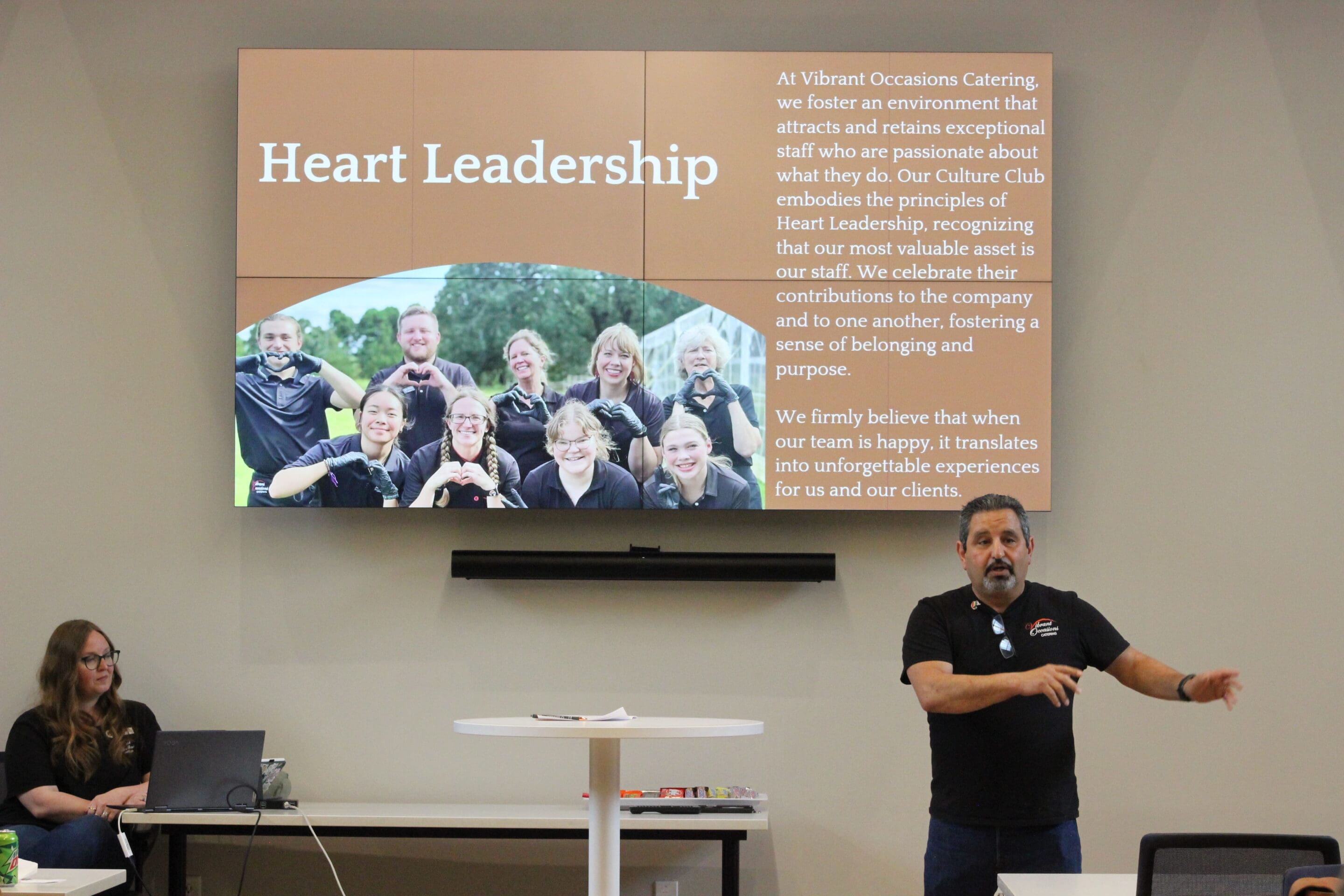A man stands and gestures while speaking in front of a screen displaying Heart Leadership with a group photo, descriptive text, and a seated woman using a laptop nearby.