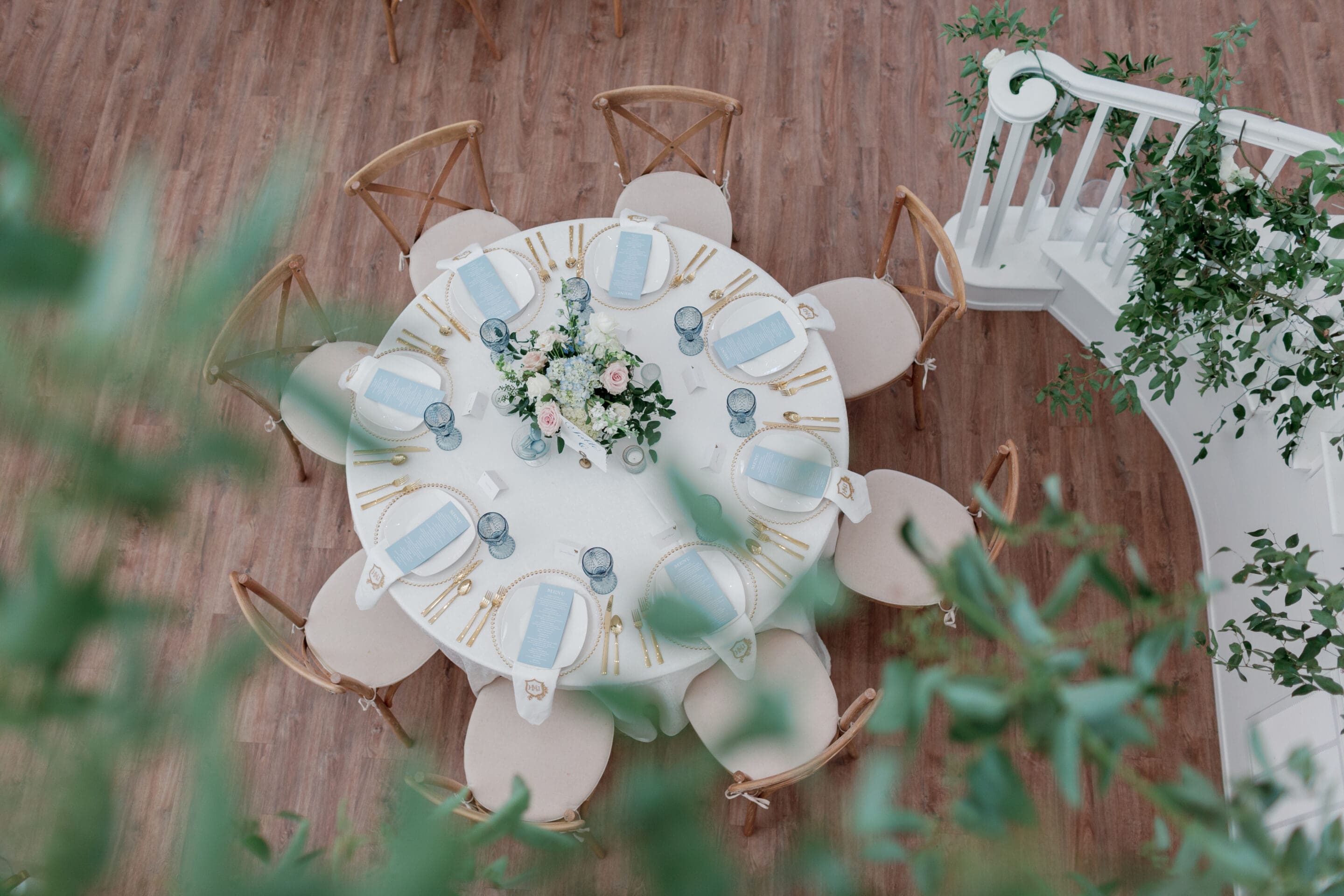 A round table set for eight with white plates, gold cutlery, blue glasses, and a floral centerpiece, viewed from above in a bright, elegant room with wood floors and greenery.