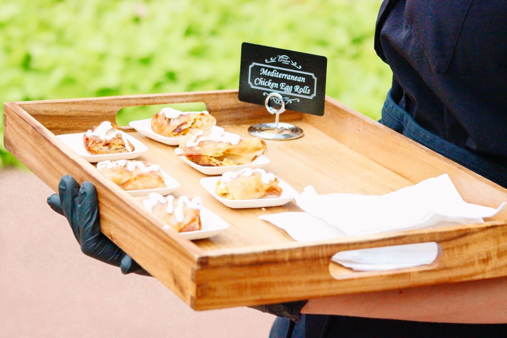 A person wearing black gloves holds a wooden tray with six small plates of Mediterranean chicken egg rolls, each topped with a drizzle of white sauce. A sign with the foods name is on the tray.