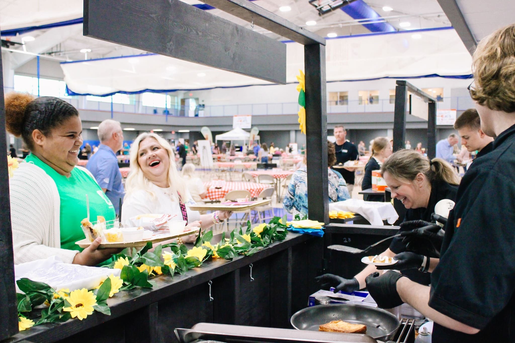 Two women smile and laugh as they receive food from workers at a booth decorated with sunflowers, inside a large, bright indoor event space filled with people and tables.