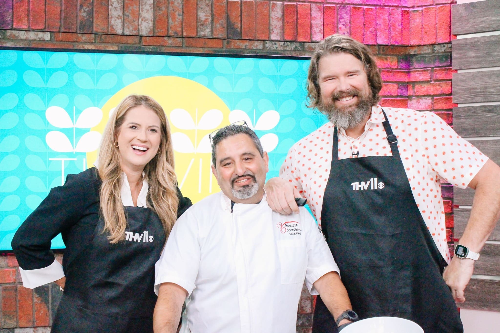 Three smiling people stand together in a kitchen studio. Two wear black THV11 aprons, and one wears a white chef jacket. They pose in front of a colorful background with a brick wall and a digital screen.