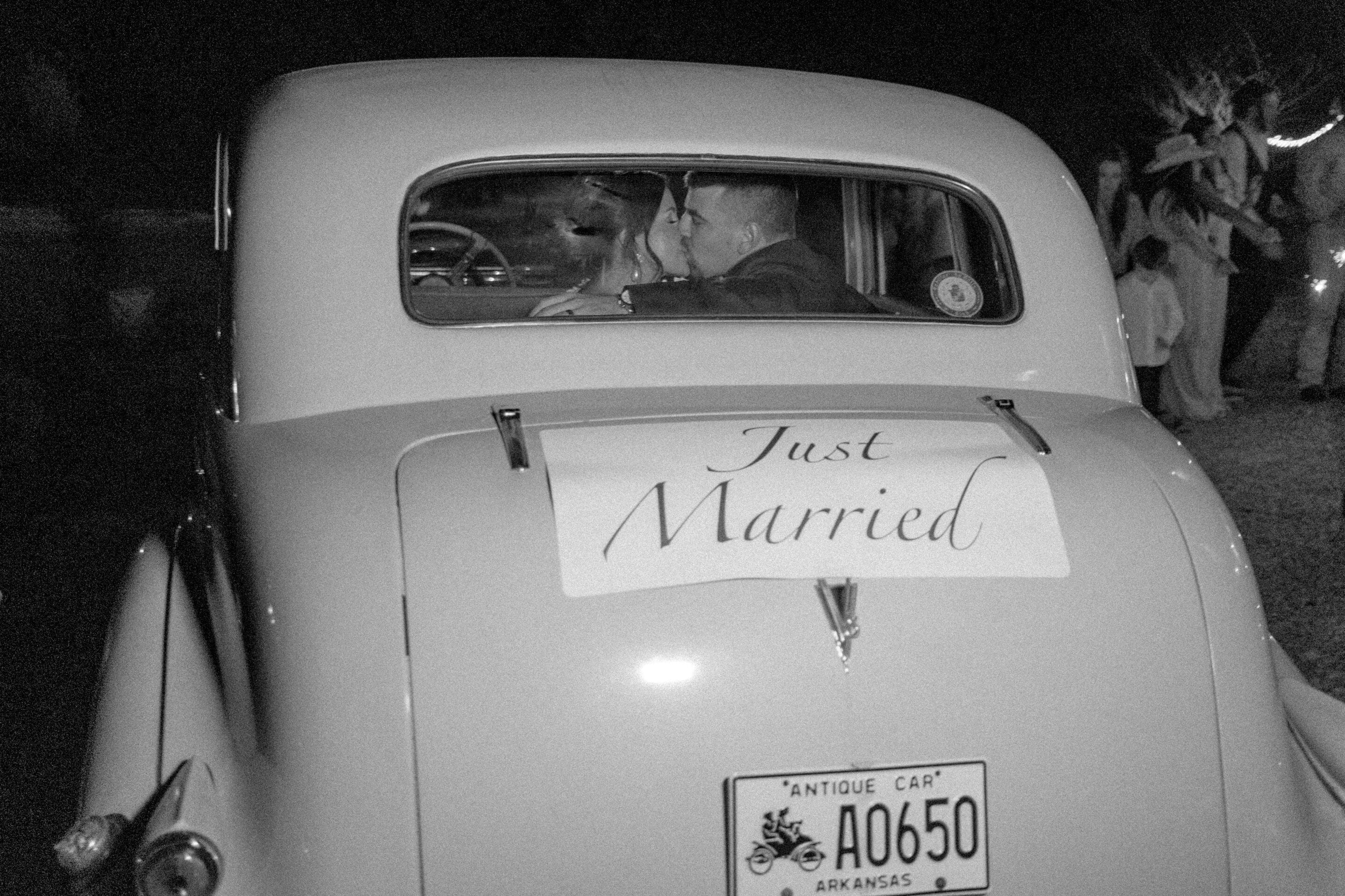 A black and white photo of a couple in a vintage car at night, seen from behind. A Just Married sign is taped to the back, and the car has an Arkansas antique license plate. People are visible in the background.