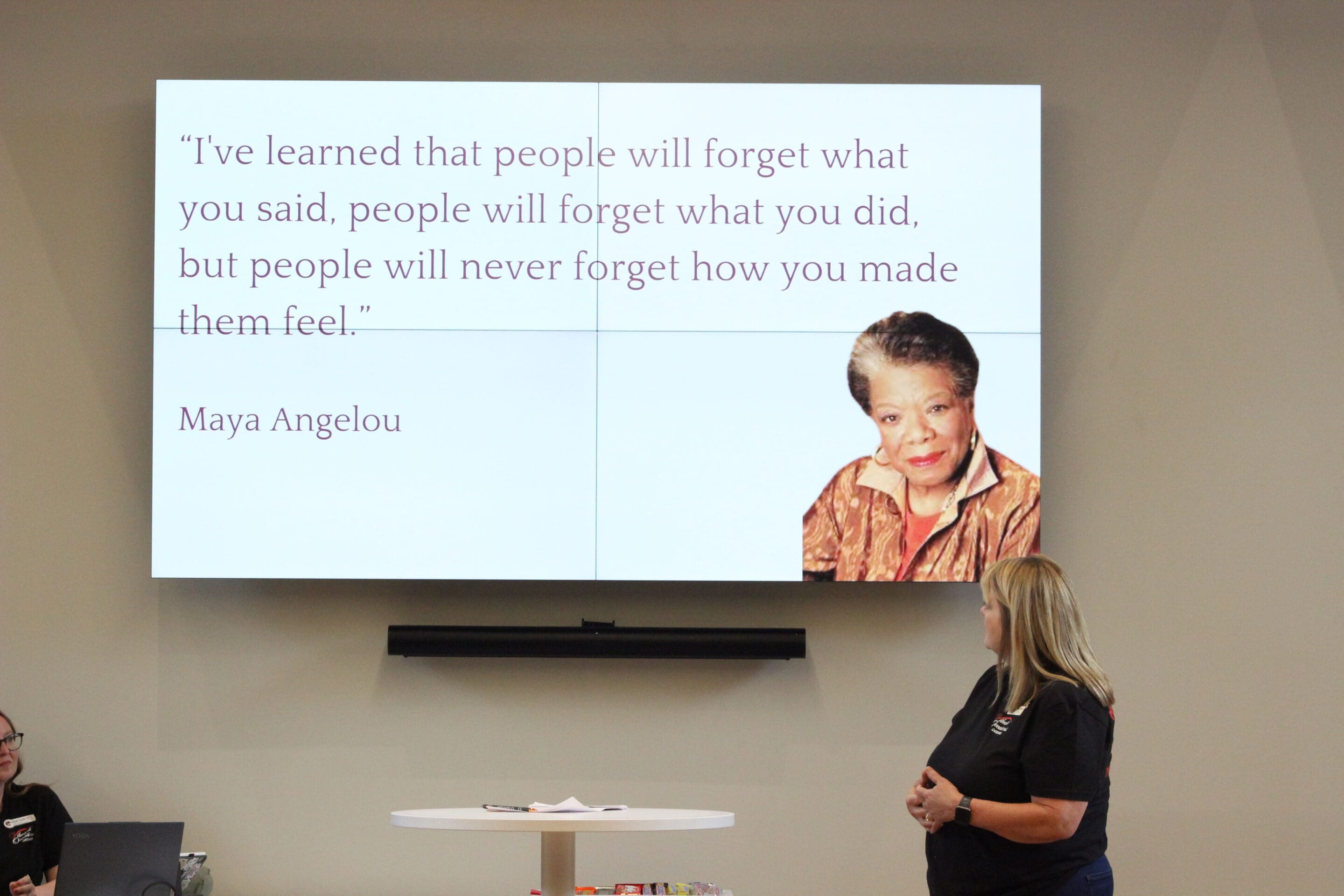 A woman stands in front of a large screen displaying a quote by Maya Angelou and her photo. The quote reads, Ive learned that people will forget what you said, people will forget what you did, but people will never forget how you made them feel.