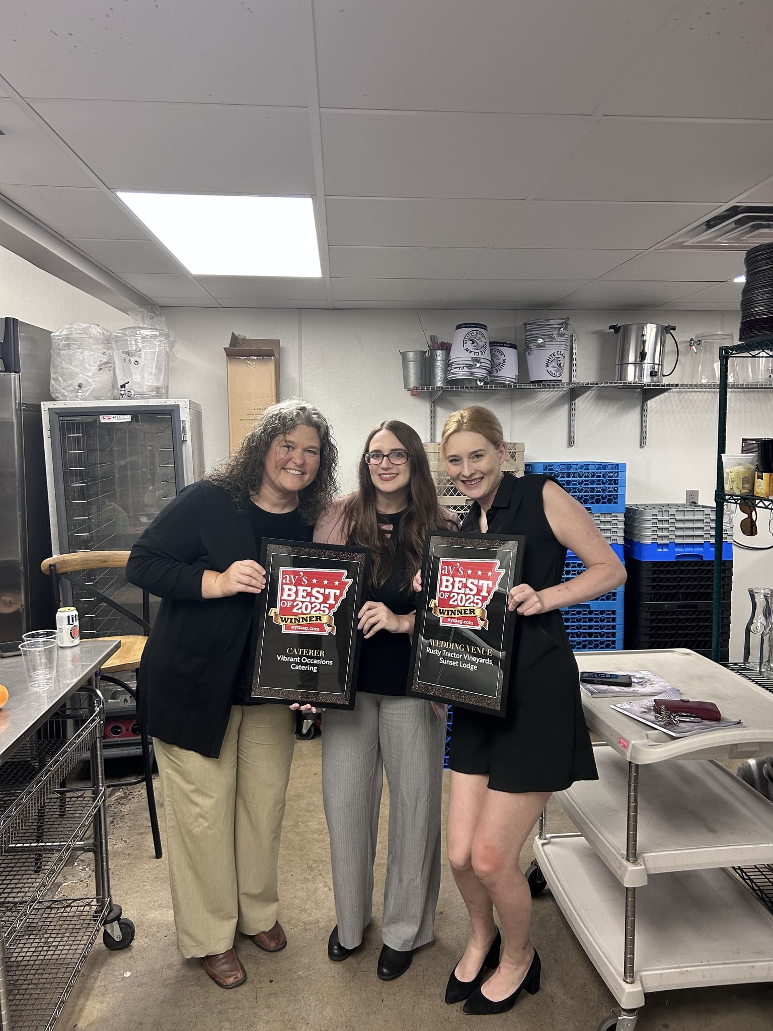 Three women standing in a commercial kitchen, smiling and holding framed Best of the Best awards. Kitchen equipment and shelves are visible in the background.