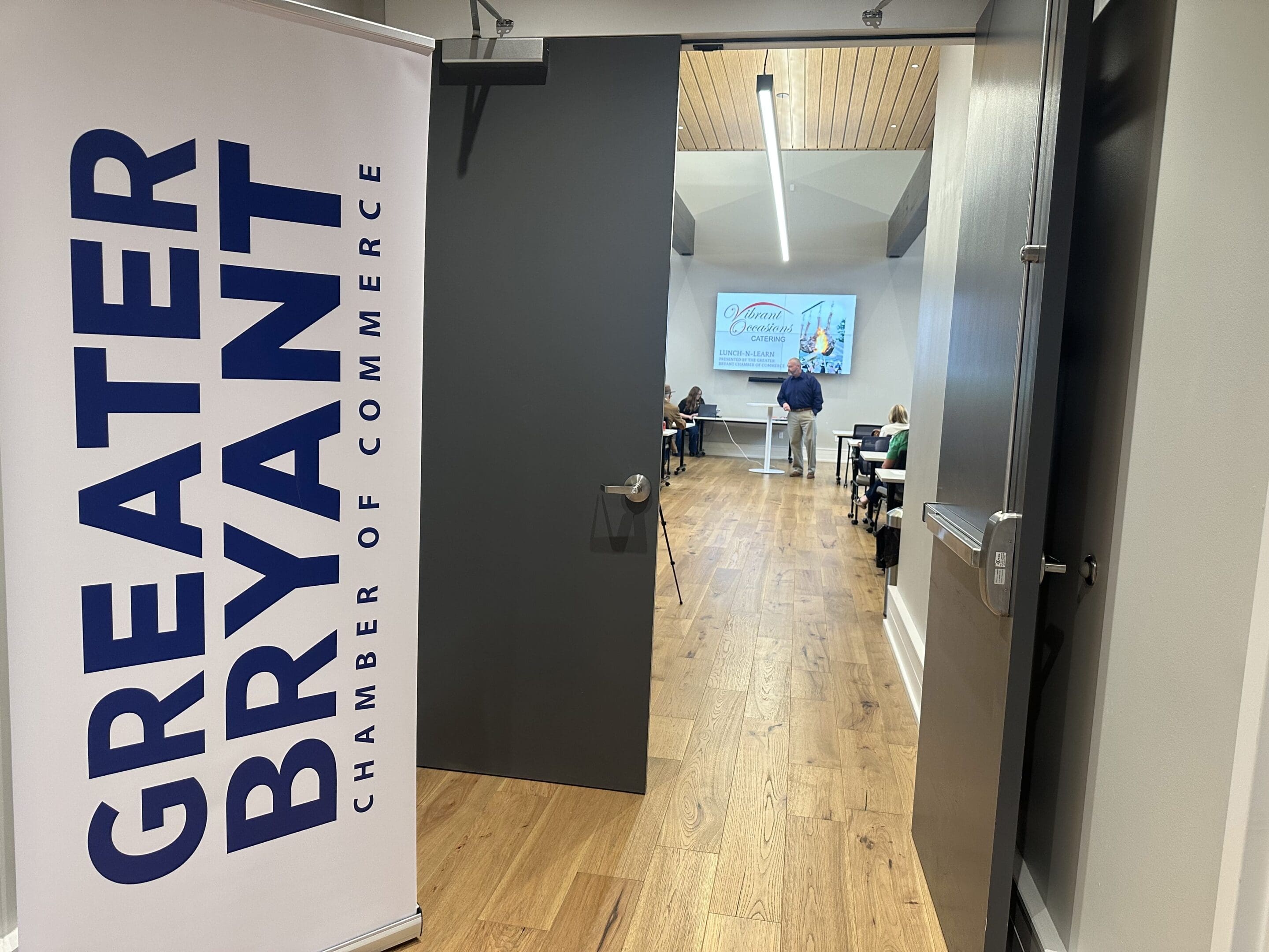 A door is open to a meeting room with people seated at tables facing a presenter and a projected screen. A sign by the door reads Greater Bryant Chamber of Commerce. The room has wood flooring and modern decor.
