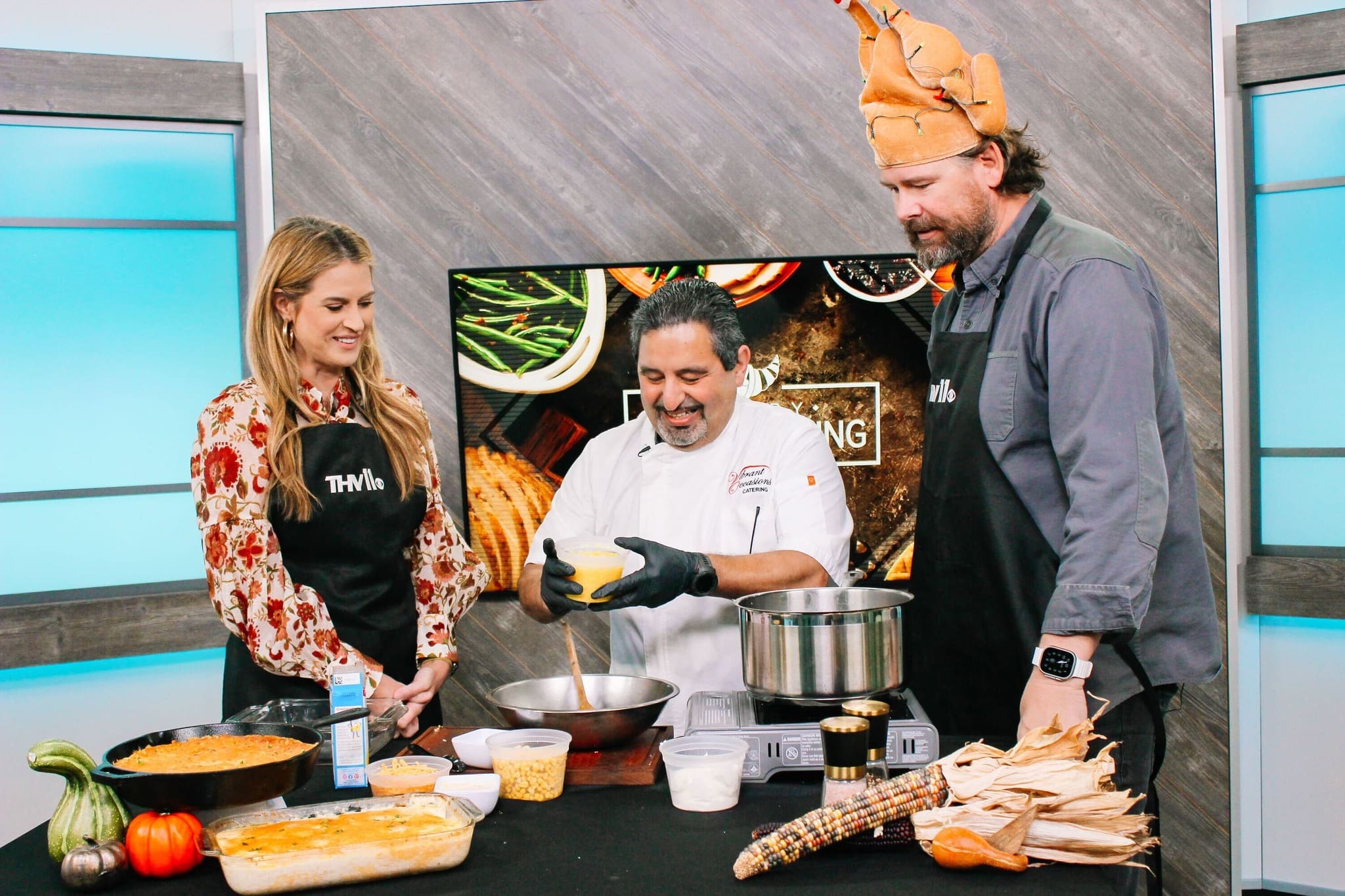 Three people stand in a kitchen studio cooking together. The chef in the center prepares food while smiling, as the man on the right wears a turkey hat. The woman on the left and the man both wear black aprons. Various foods are on the counter.