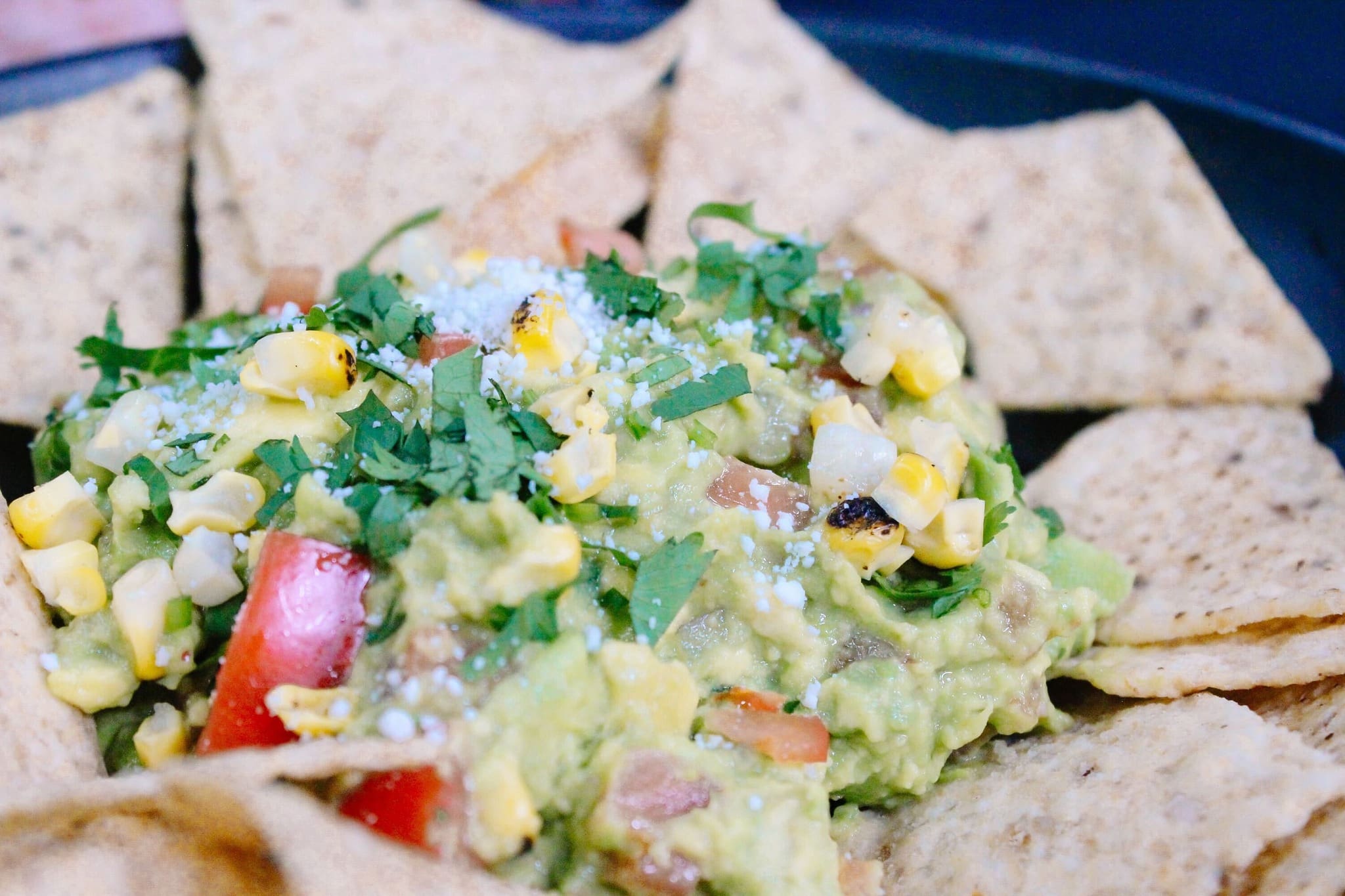 A fun guacamole bar features a plate of tortilla chips arranged around a mound of guacamole topped with corn, diced tomatoes, cilantro, and crumbled cheese.