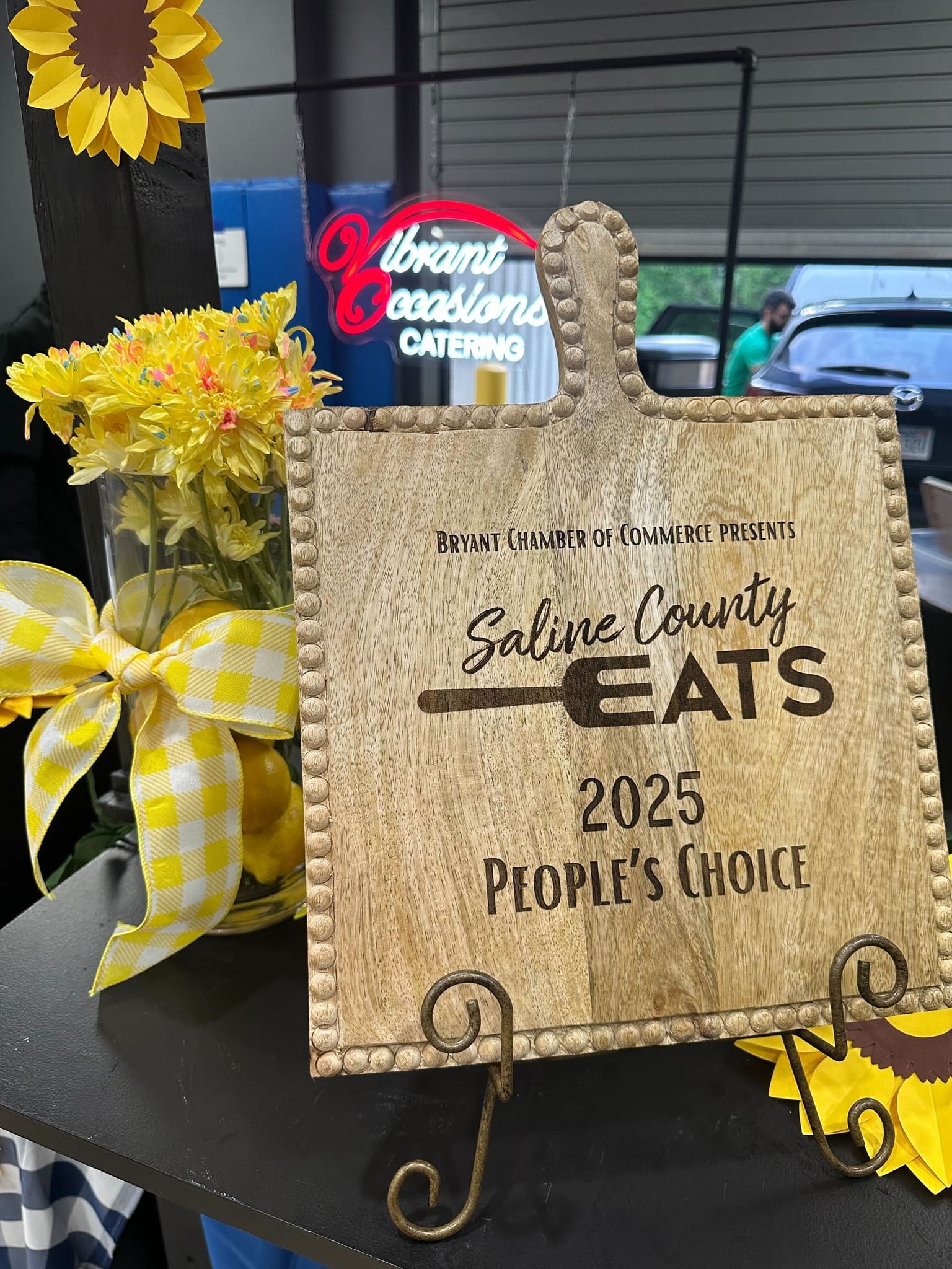 A wooden plaque reads “Saline County Eats 2025 People’s Choice” from the Bryant Chamber of Commerce. It’s displayed on a stand next to a mason jar of yellow flowers with a yellow plaid bow, and a sunflower decoration.