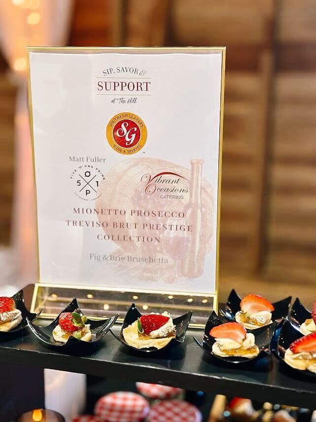 A display of hors d’oeuvres on black plates sits in front of a sign promoting a catering event featuring bruschetta and Mionetto Prosecco, with decorative wine graphics in the background.