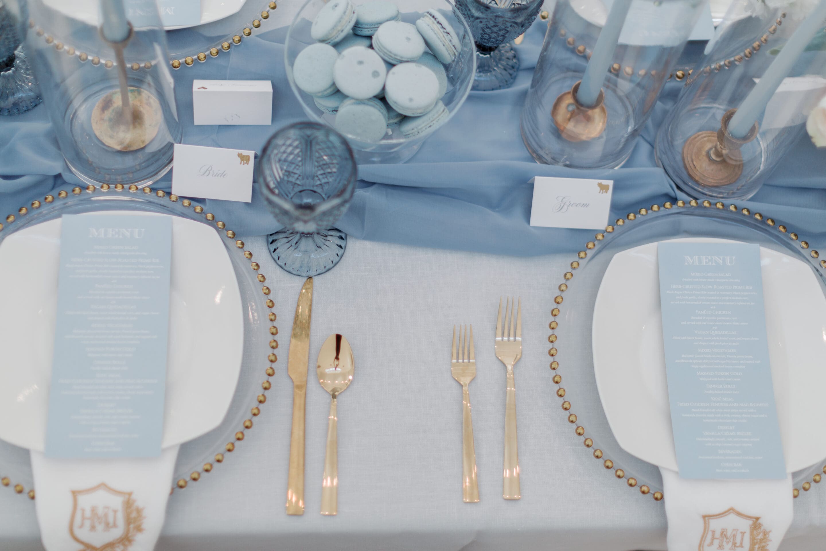 Elegant table setting with gold cutlery, white plates, blue menus, blue glassware, and blue macarons on a centerpiece. Blue and white cards mark places at a table with a pale blue tablecloth and blue candles.