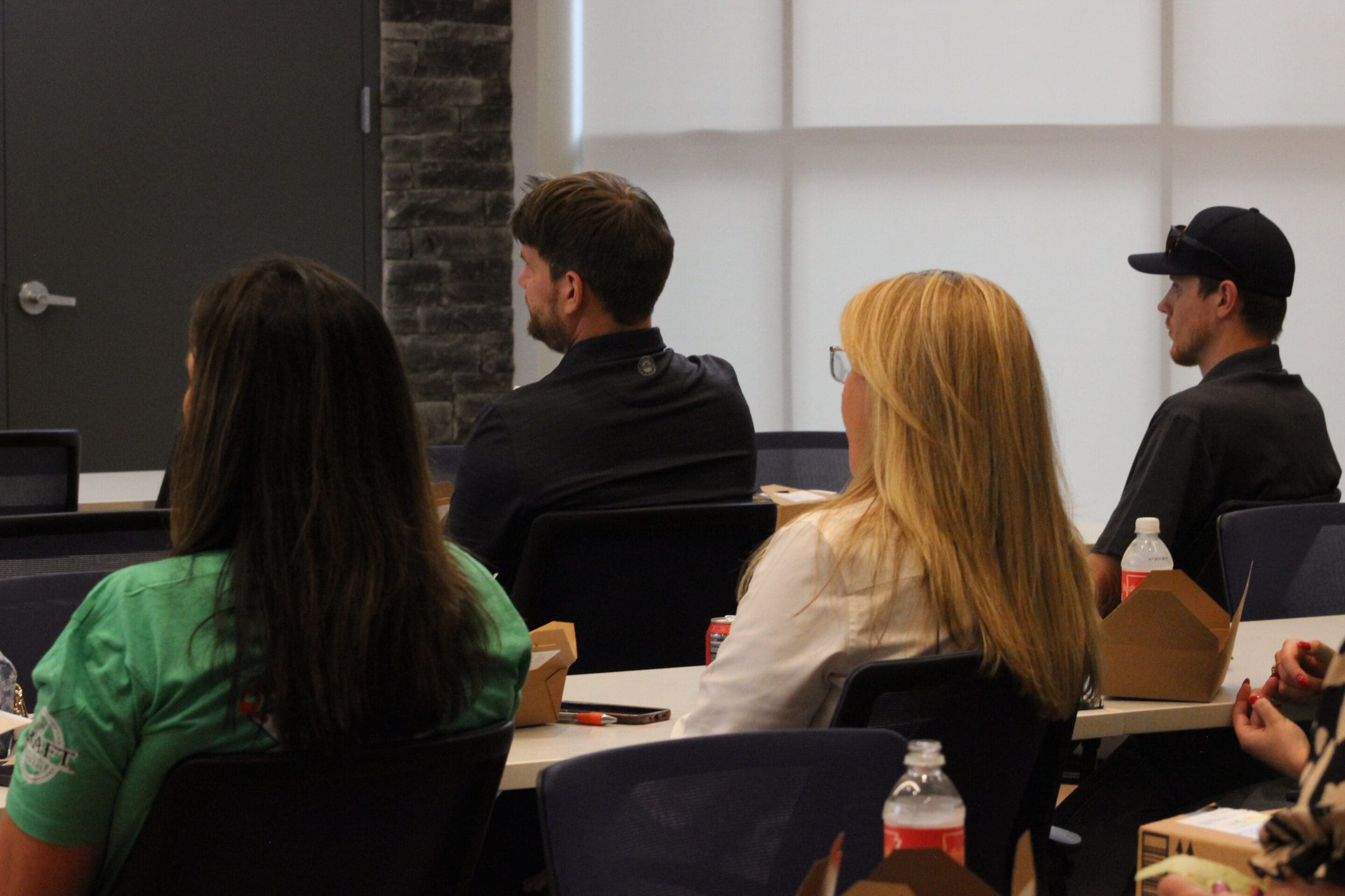 Several people sit at tables in a classroom or meeting room, facing forward. Lunch boxes and bottles of water are in front of them. The background includes a brick wall and large windows with shades.