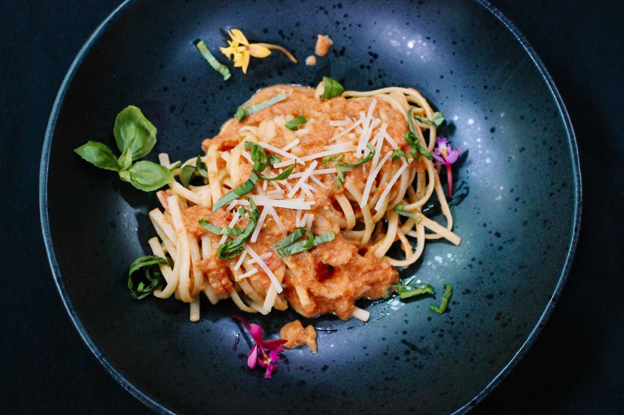 A black plate with spaghetti topped with tomato sauce, grated cheese, chopped basil, and garnished with fresh basil leaves and small colorful edible flowers.