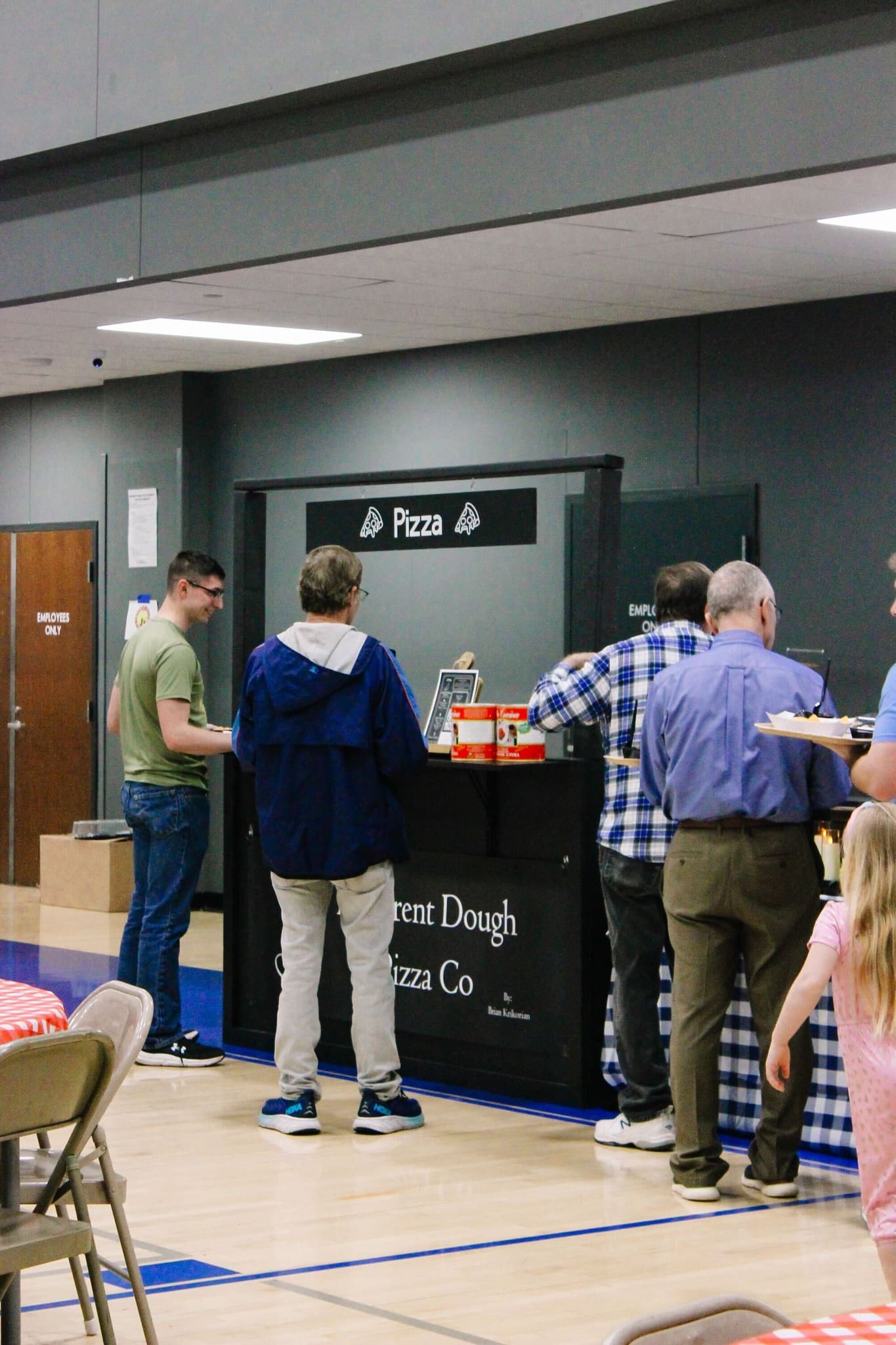 People stand in line at a pizza stall inside a gymnasium, waiting to be served. The stall has a sign that says “Pizza.” Nearby tables are covered with blue checkered cloths. A young girl stands in the foreground.