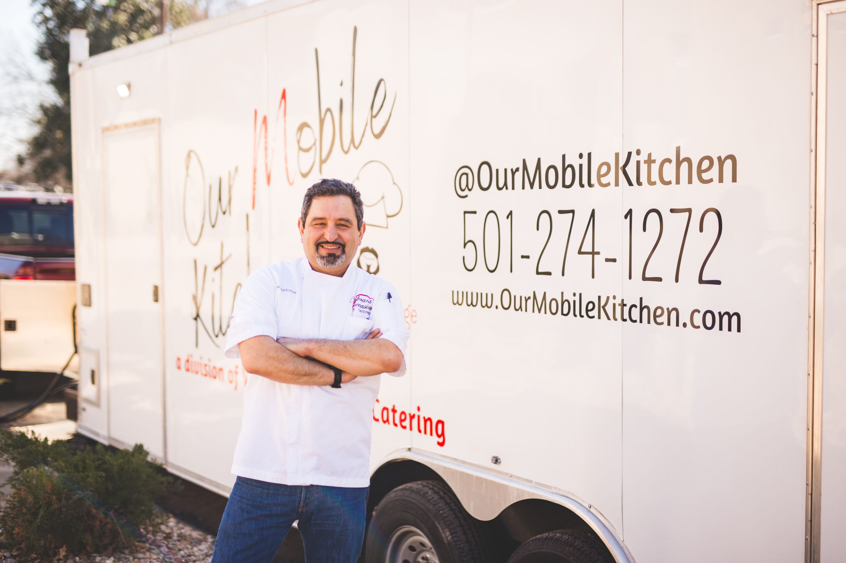 A man in a white chefs coat stands smiling with his arms crossed in front of a food truck labeled Our Mobile Kitchen with contact information displayed on the side.