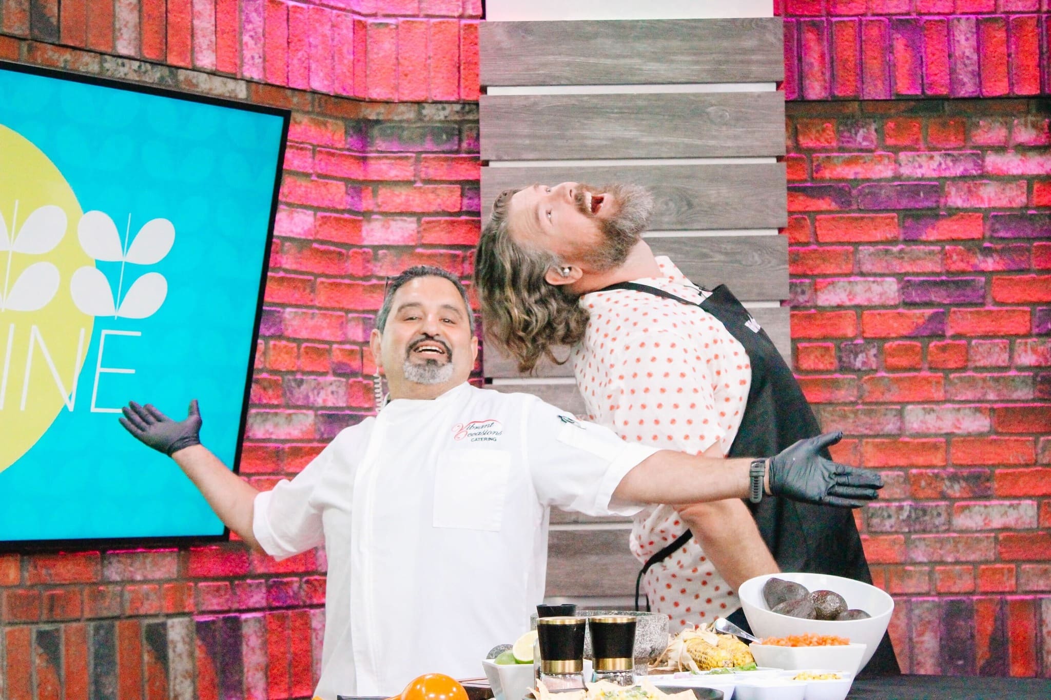 Two chefs stand back-to-back in a playful pose, arms outstretched and smiling widely, ready to create at a fun guacamole bar. The colorful kitchen features a red brick wall, bright decor, and a counter with fresh ingredients in the foreground.