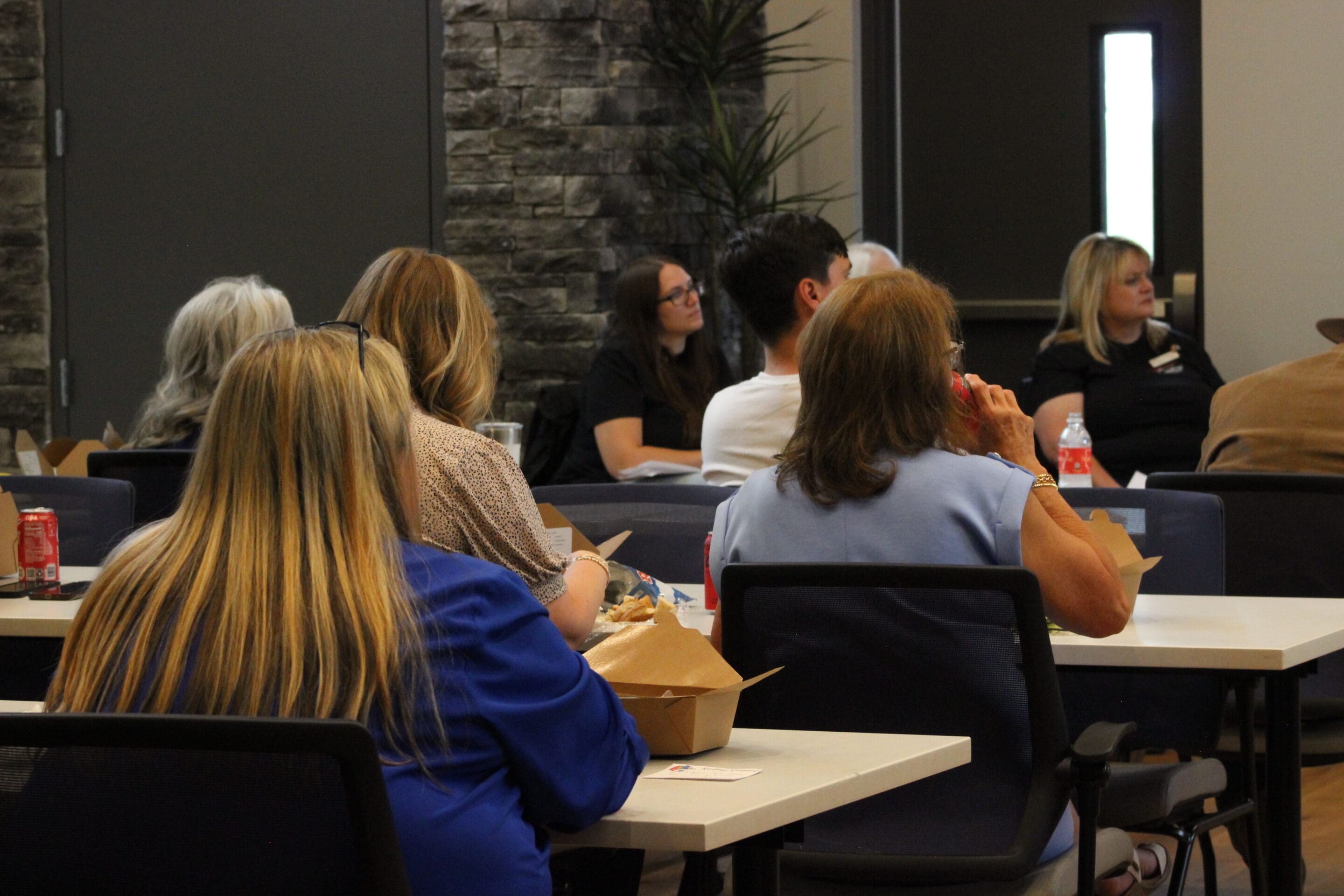 People sit at tables in a conference room, listening attentively. Some are eating from takeout boxes, with drinks like soda on the tables. The room has a modern design, with stone walls and plants visible.