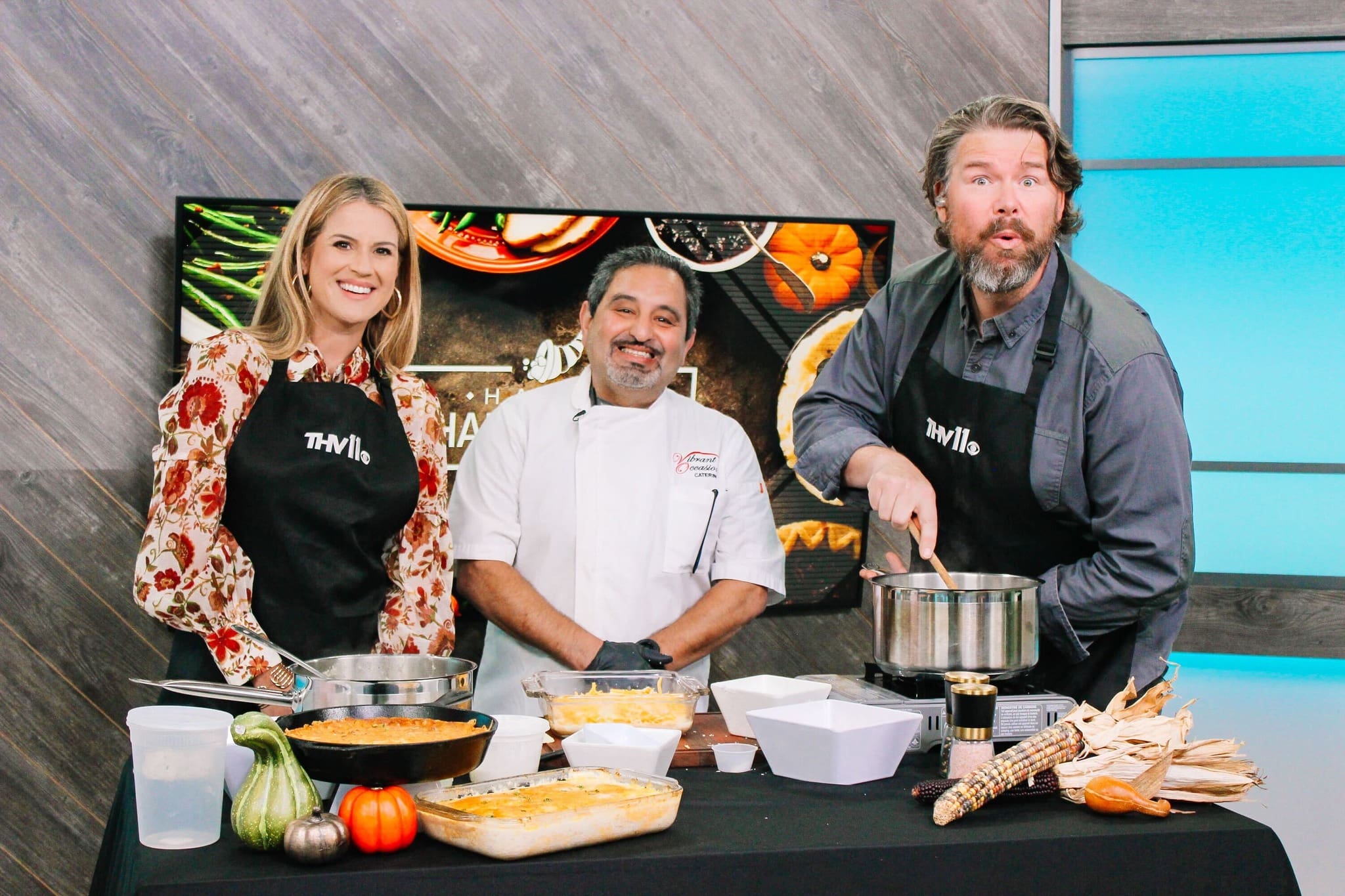 Three people stand behind a table with prepared dishes and cooking utensils. One stirs a pot, another stands smiling in a chefs coat, and the third smiles at the camera. A TV screen and fall decor are in the background.