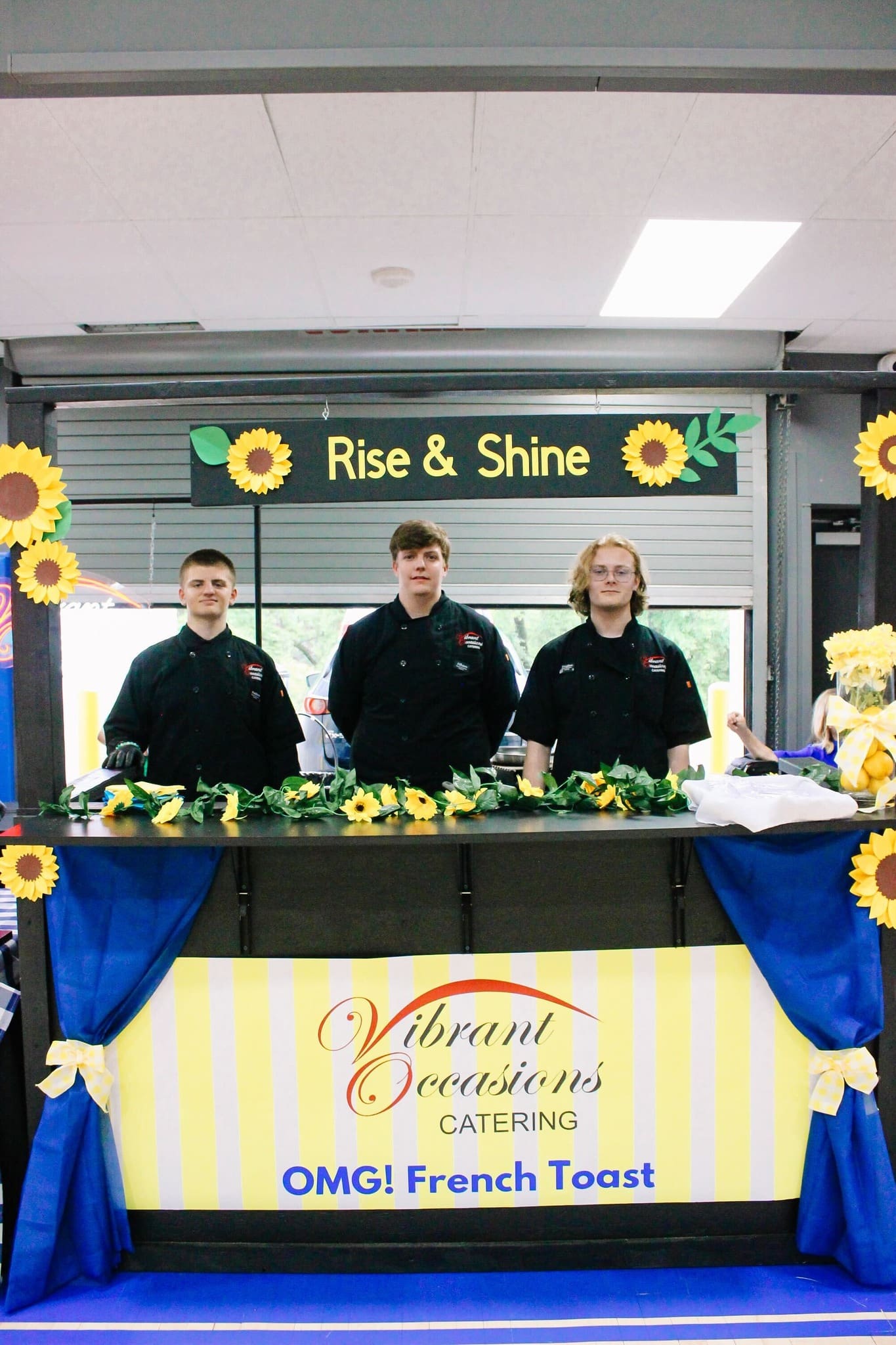 Three people in black uniforms stand behind a decorated booth with sunflowers and a sign reading “Rise & Shine.” The counter displays “Vibrant Occasions Catering: OMG! French Toast” on a yellow and white striped banner.
