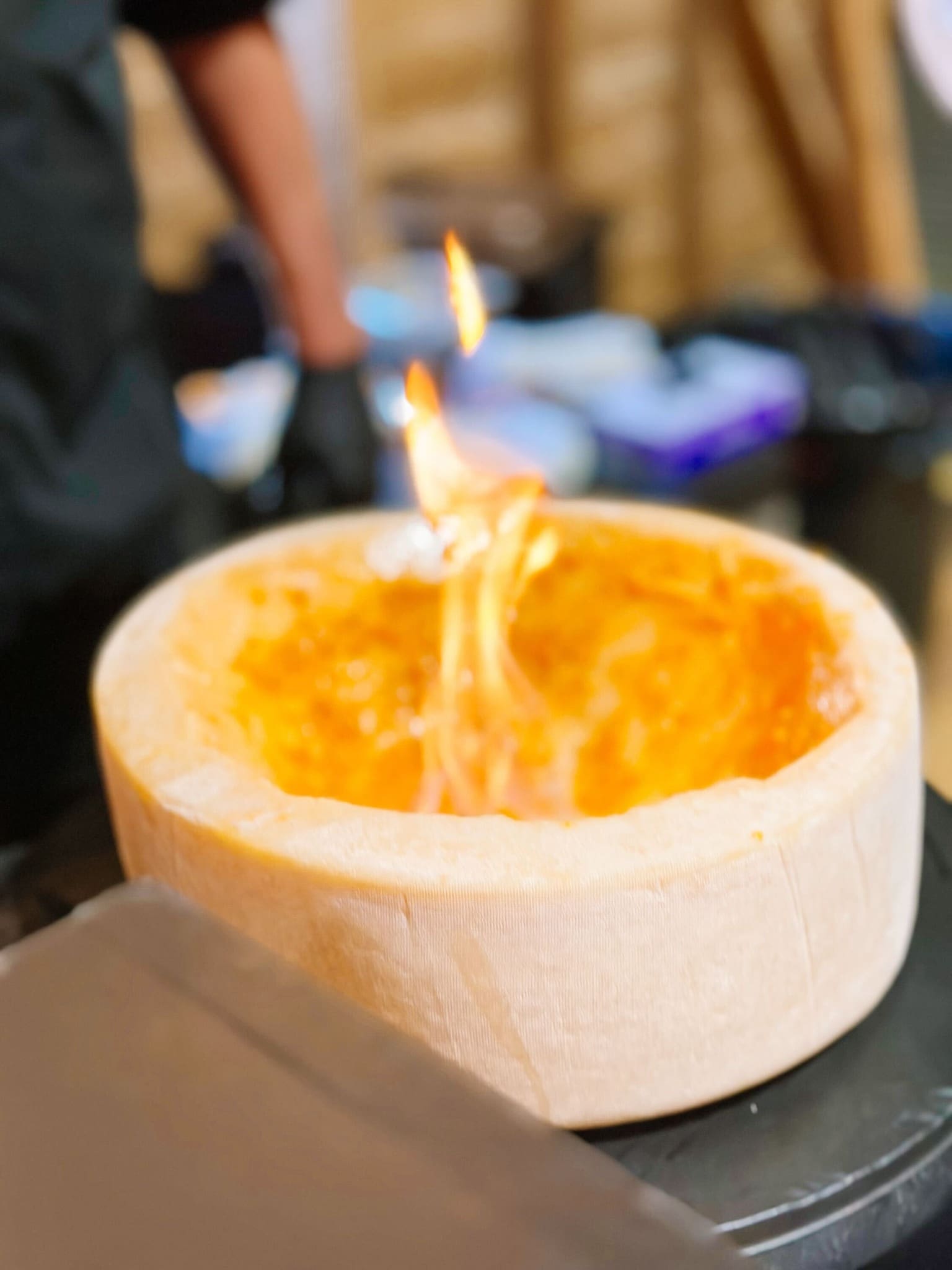 A large wheel of cheese with a melted, golden interior is set aflame on top, creating a dramatic fiery effect. The background is blurred, drawing focus to the flaming cheese.