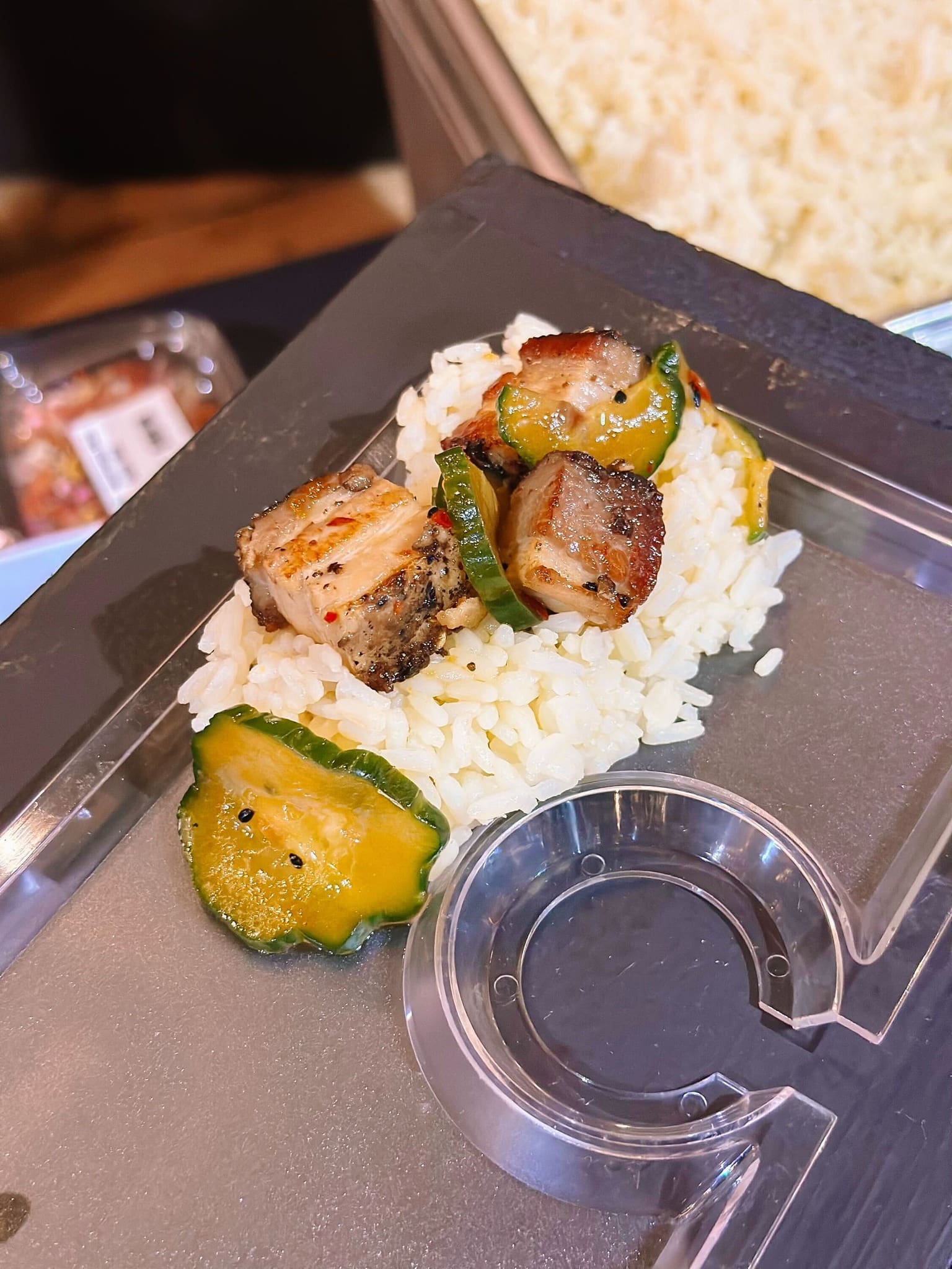 A serving of white rice topped with grilled pork belly slices and pickled cucumber pieces on a clear plastic plate.