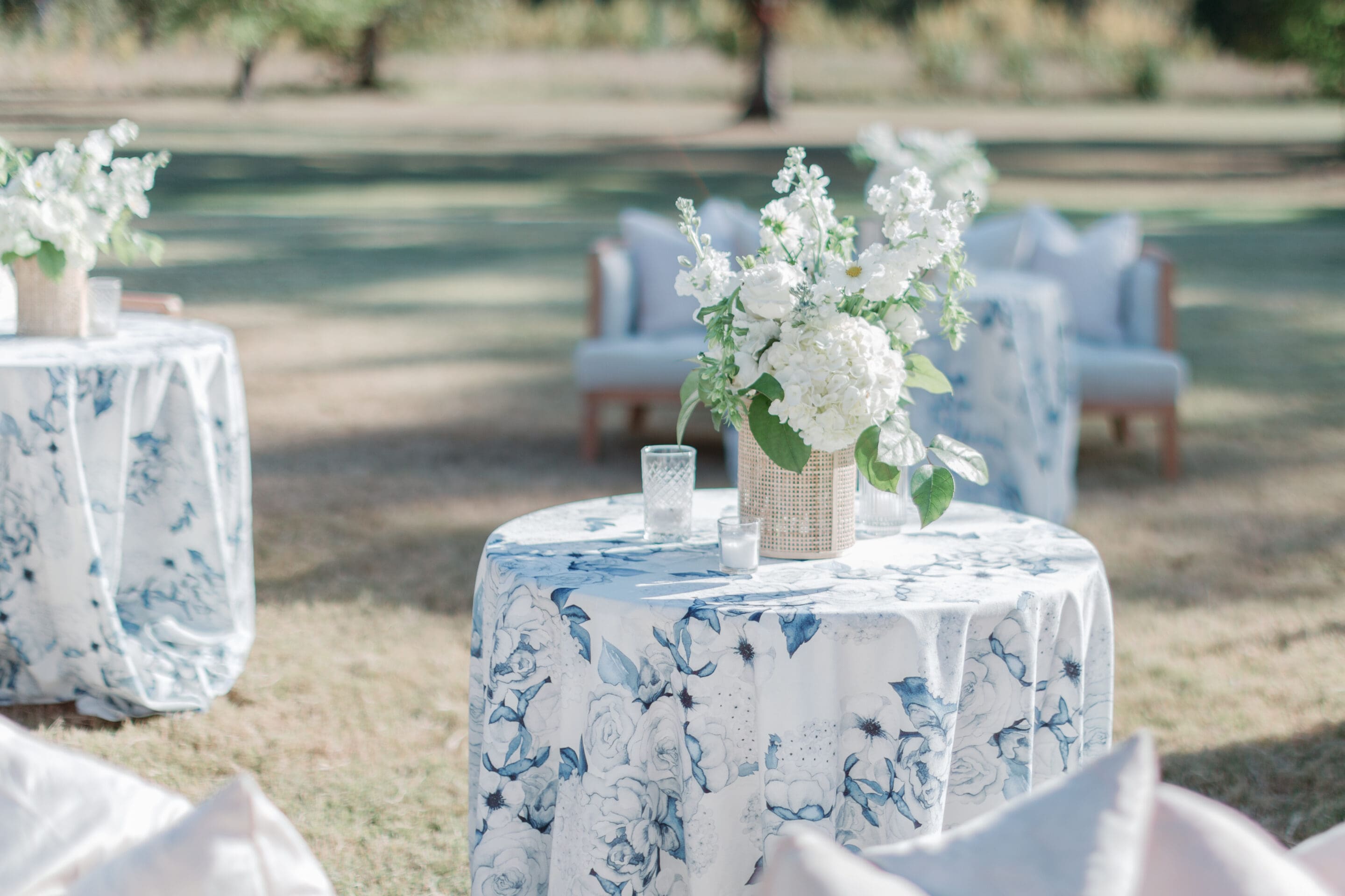 A round table with a blue and white floral tablecloth holds a wicker vase of white flowers and a glass candle holder, set outdoors on grass with armchairs and more decorated tables in the background.