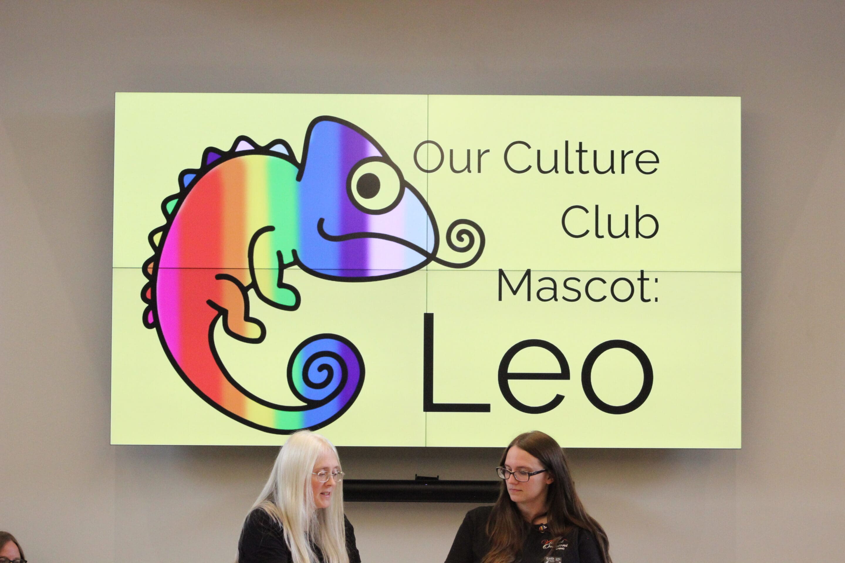 A large screen displays a colorful chameleon with the text Our Culture Club Mascot: Leo. Two women with long hair sit in front of the screen, one with white hair and one with brown hair.