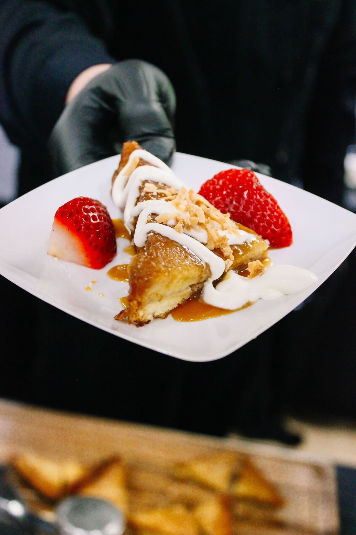 A person wearing a black glove holds a plate with a slice of dessert topped with white icing, caramel sauce, chopped nuts, and served with two strawberry halves.