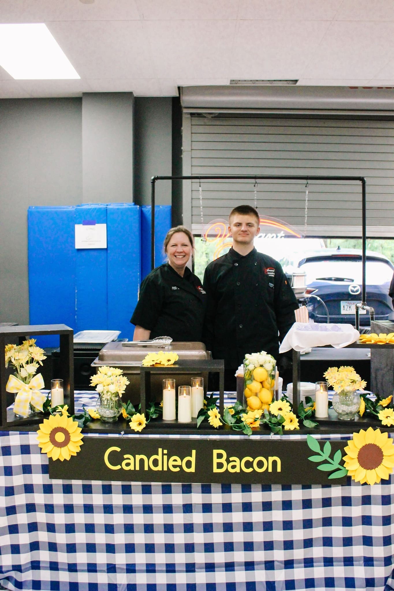 Two people in black chef coats stand behind a “Candied Bacon” booth decorated with sunflowers, lemons, candles, and a blue-checkered tablecloth. Plates of food are set out on the table.