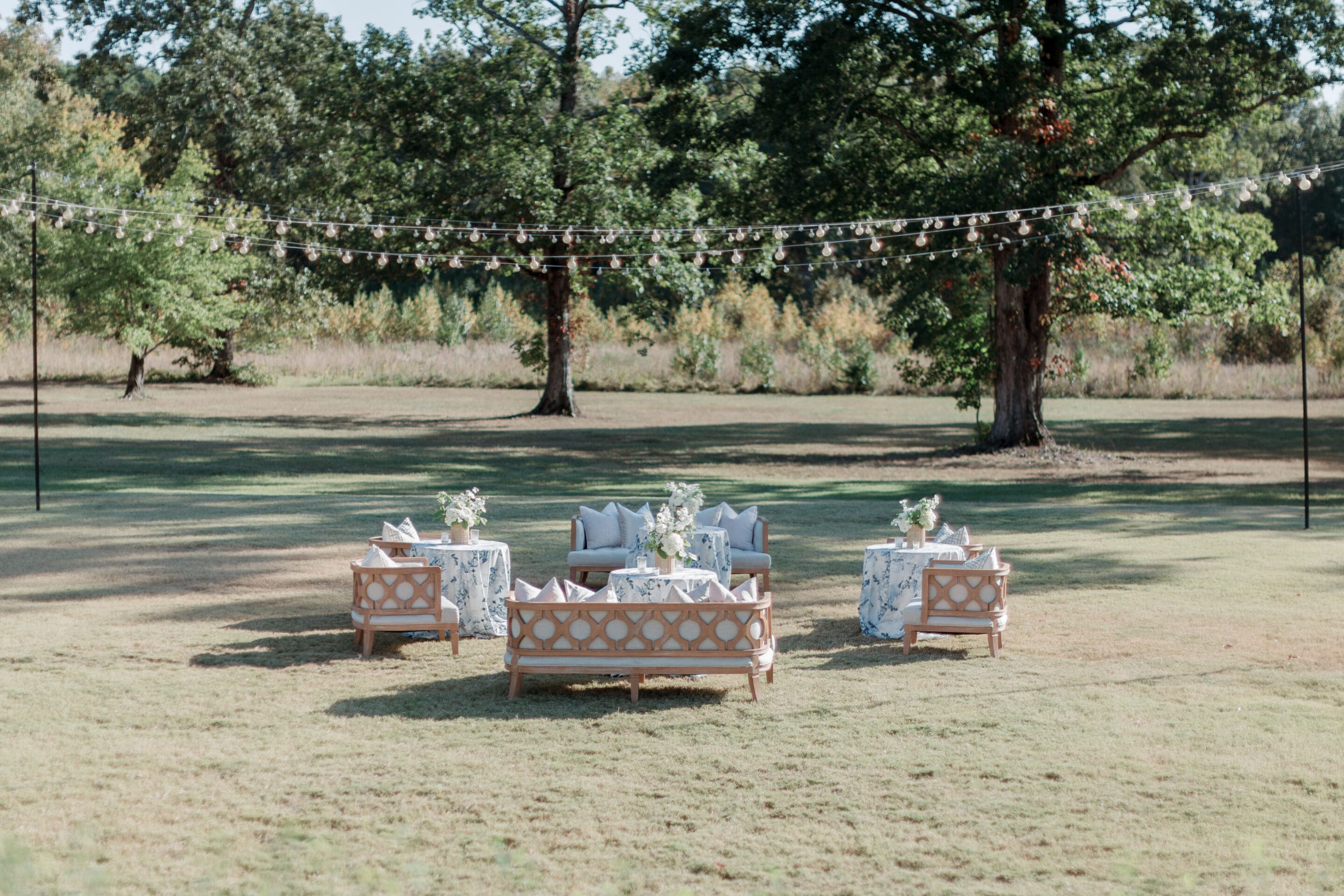 Four sofas with patterned blue and white tablecloth-covered tables form a lounge area on a grassy lawn, surrounded by trees and string lights, perfect for an Elegant Spring Wedding at The Grandeur House in Little Rock.