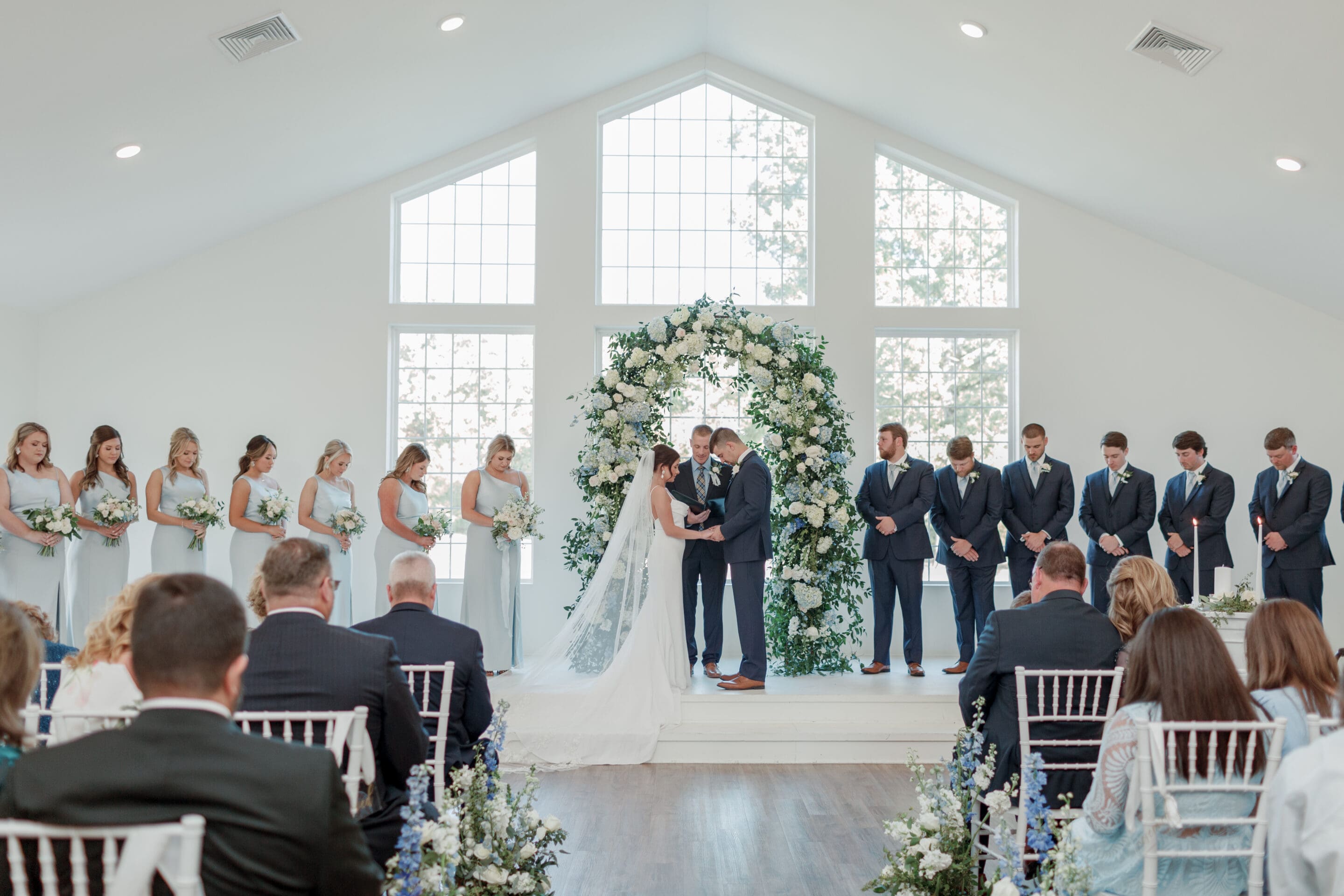 A bride and groom stand at the altar holding hands, surrounded by their wedding party in a bright, flower-adorned chapel with large windows and seated guests watching the ceremony.