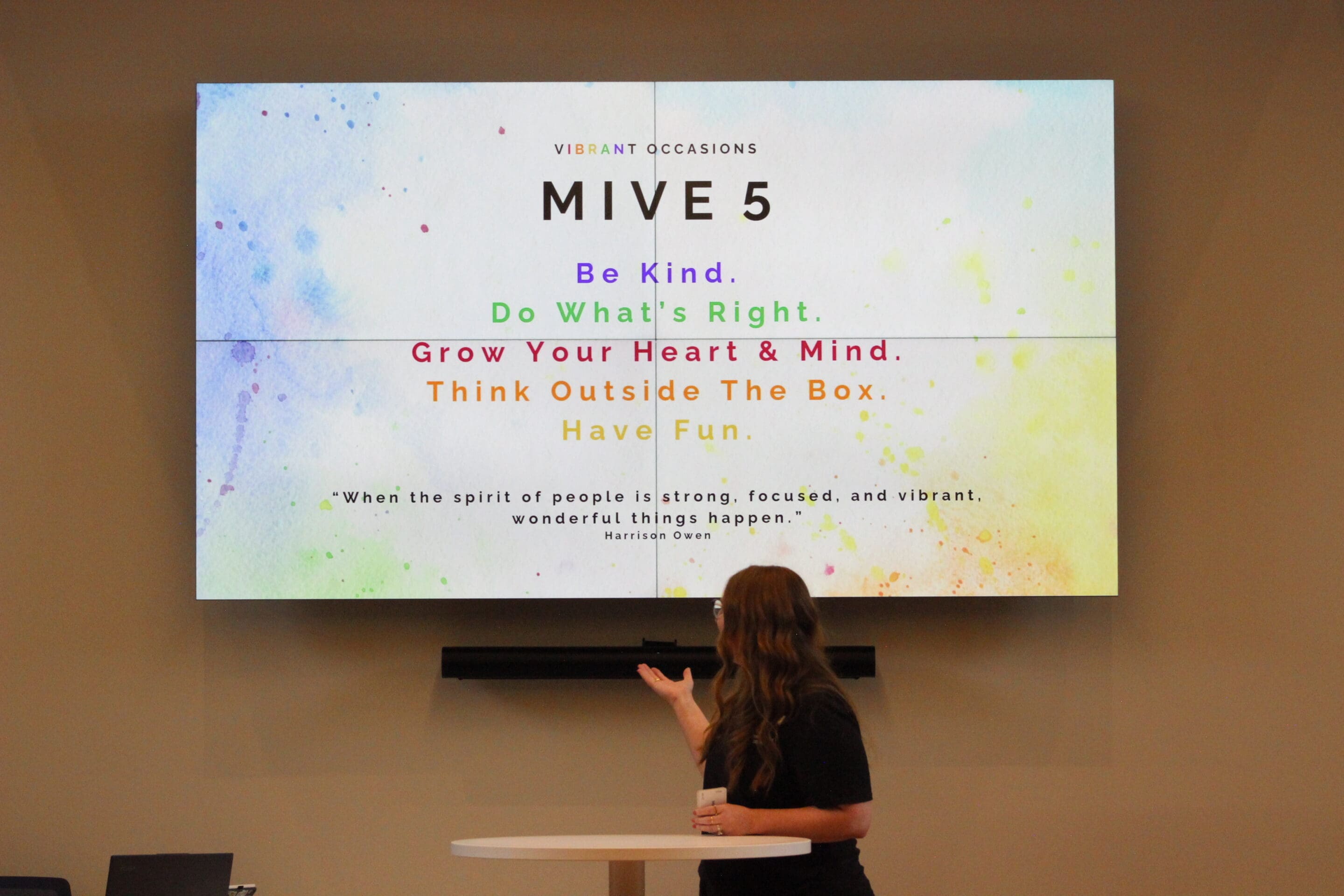 A woman gestures toward a colorful presentation slide displaying MIVE 5 with the phrases: Be Kind, Do Whats Right, Grow Your Heart & Mind, Think Outside The Box, and Have Fun, along with a motivational quote.