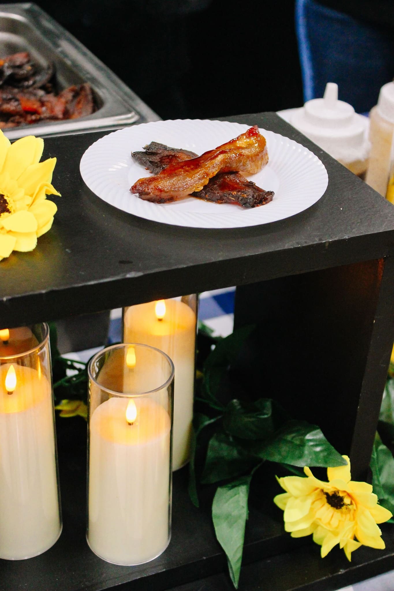A white plate with several pieces of cooked bacon sits on a black shelf decorated with artificial sunflowers, green leaves, and lit pillar candles in glass holders. Squeeze bottles are visible in the background.