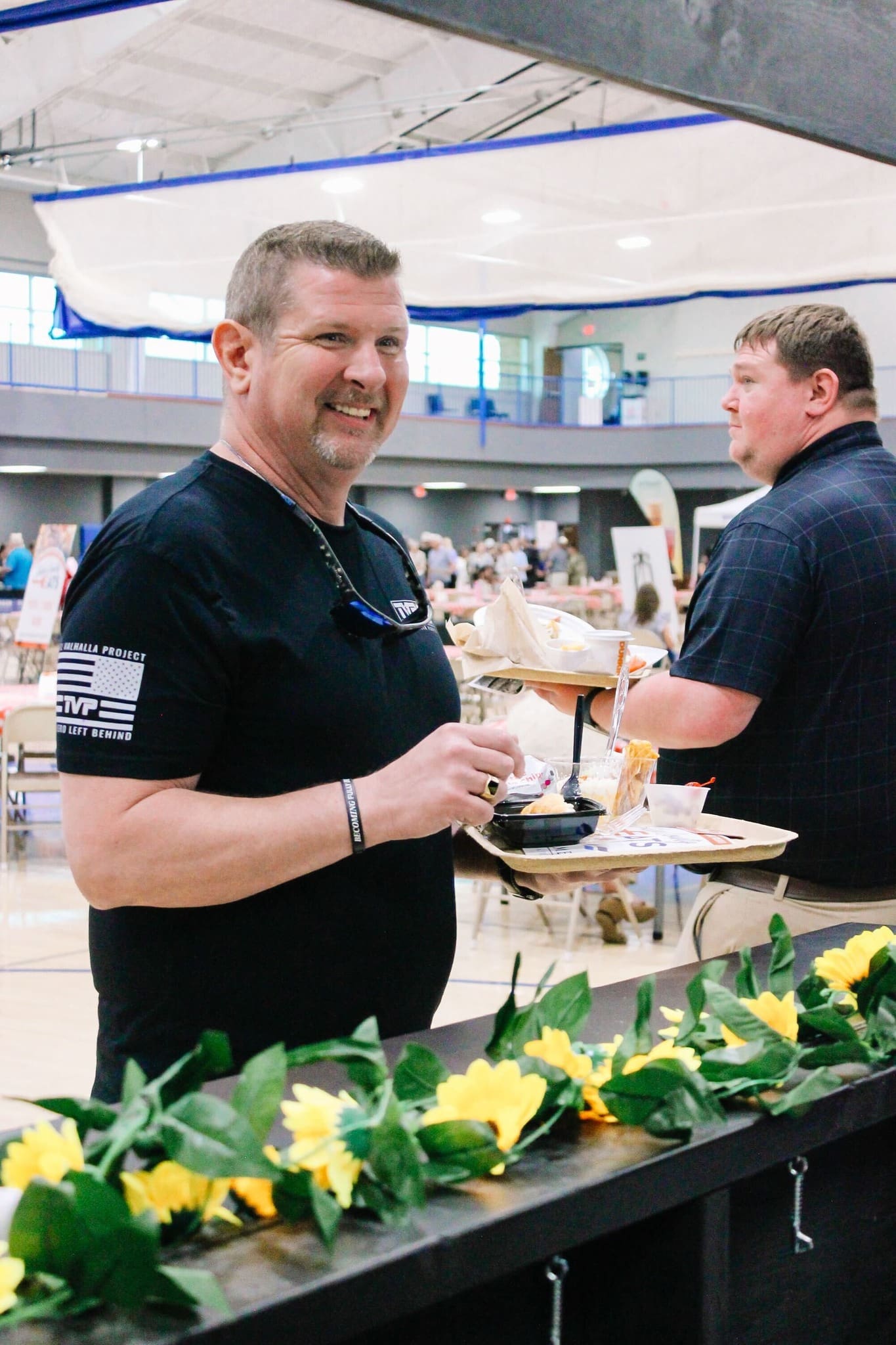 A smiling man in a black t-shirt holds a plate of food at an indoor event. Another man stands behind him, and sunflowers decorate the foreground. The background shows tables and people in a gymnasium setting.