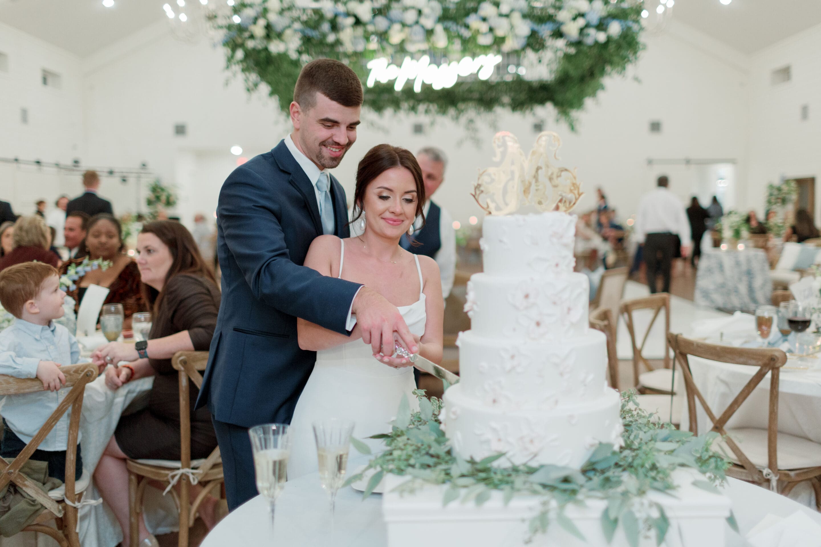 A bride and groom, smiling, cut a white, three-tiered wedding cake together at a decorated reception with guests seated in the background and greenery overhead.