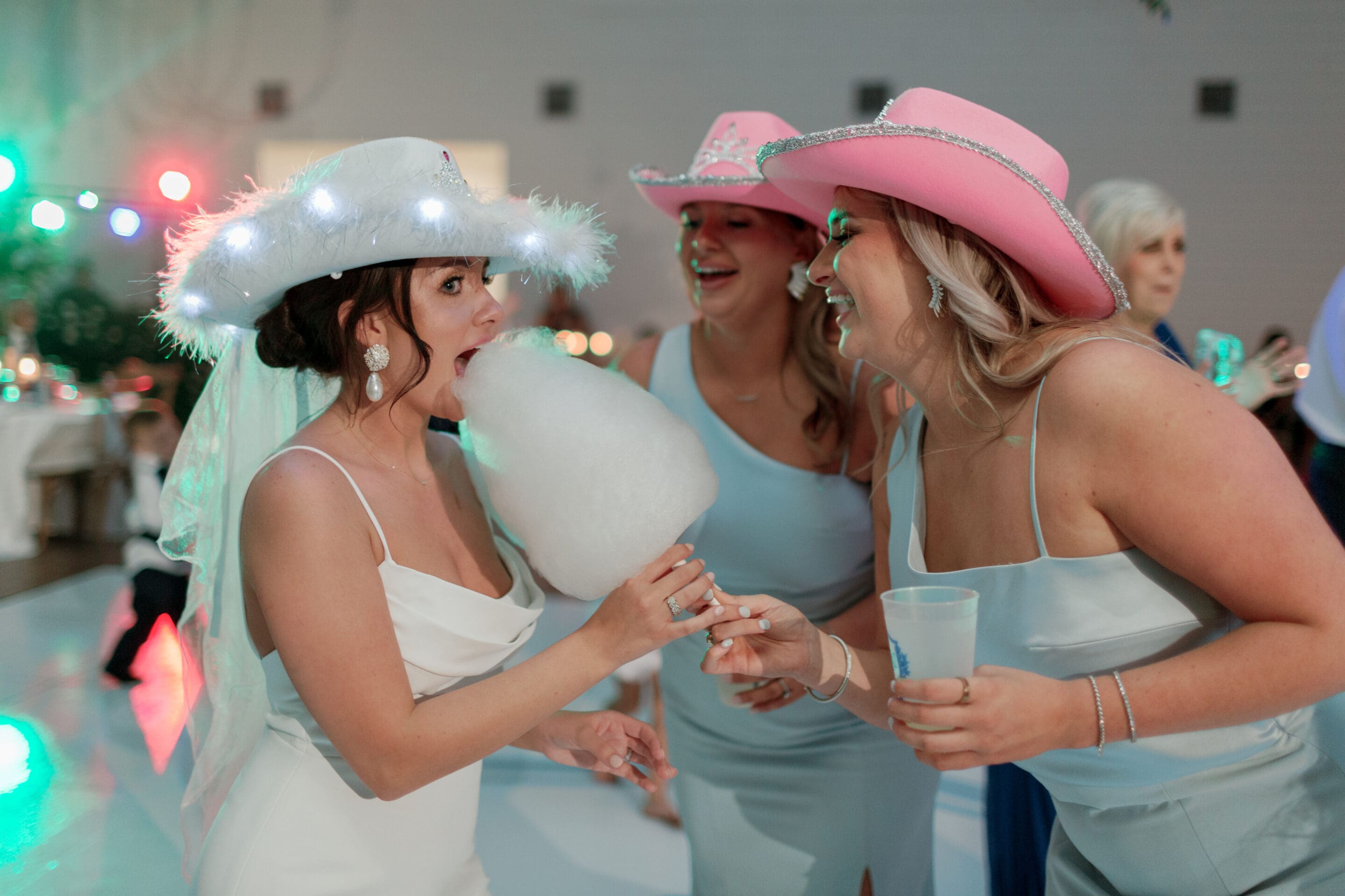 A bride in a white dress and light-up hat eats cotton candy while laughing with two bridesmaids in pink cowboy hats and light blue dresses at a lively indoor celebration.