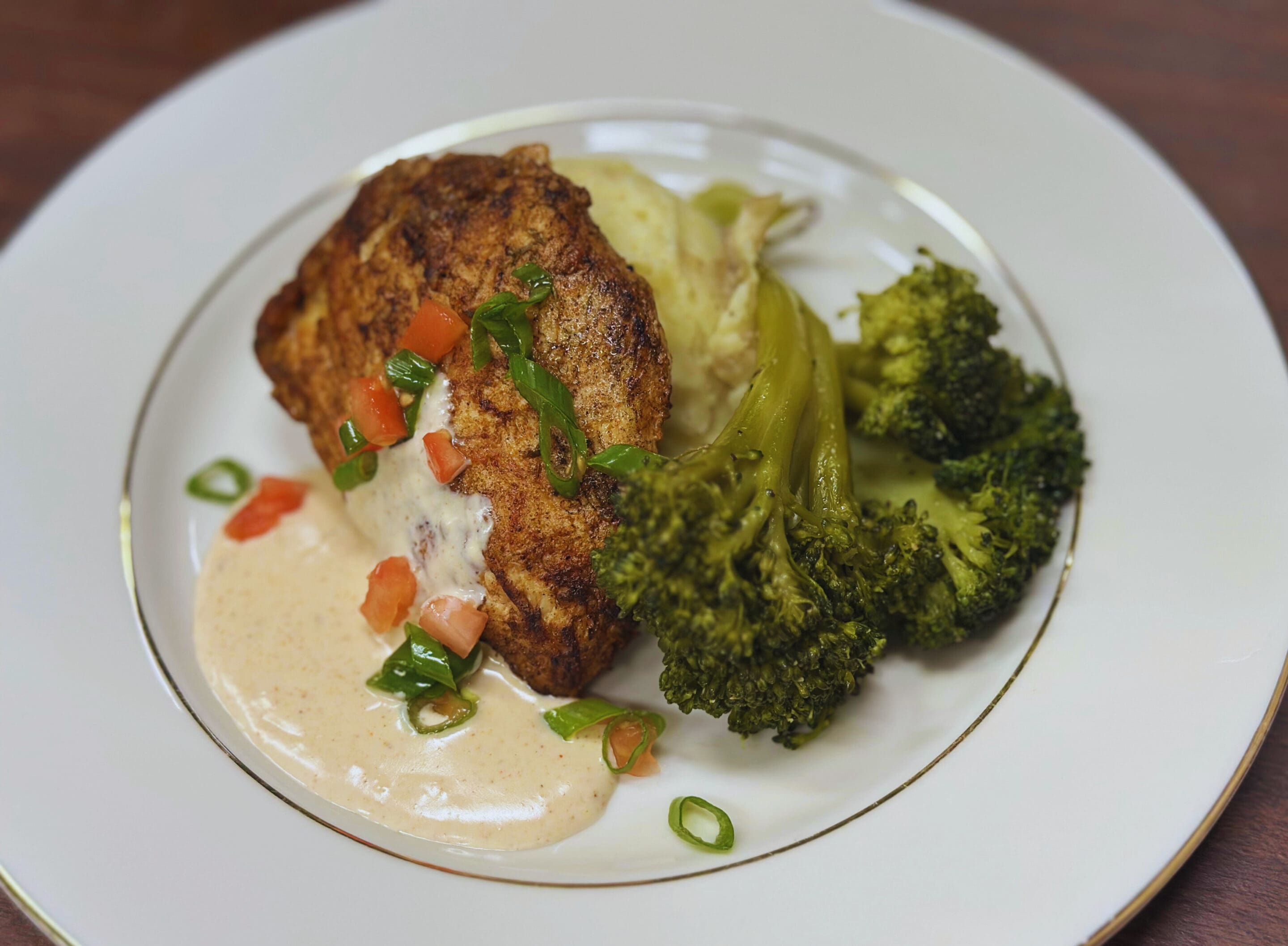 A plated meal featuring a seasoned chicken breast with creamy sauce and diced tomatoes, served alongside mashed potatoes and steamed broccoli on a white plate.