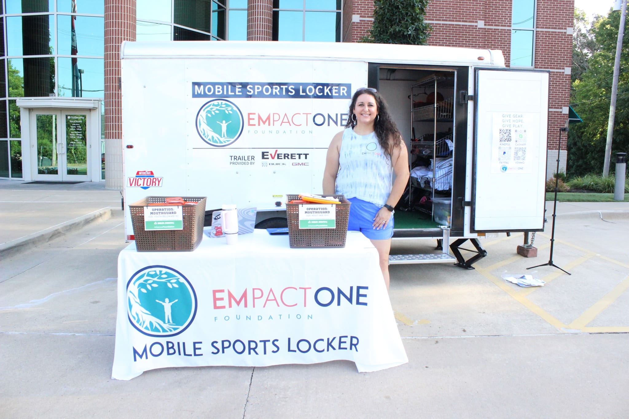 A woman stands beside a trailer labeled Mobile Sports Locker and Empact One Foundation. Two tables in front hold baskets with donated sports equipment and flyers. The scene is outdoors near a modern building.