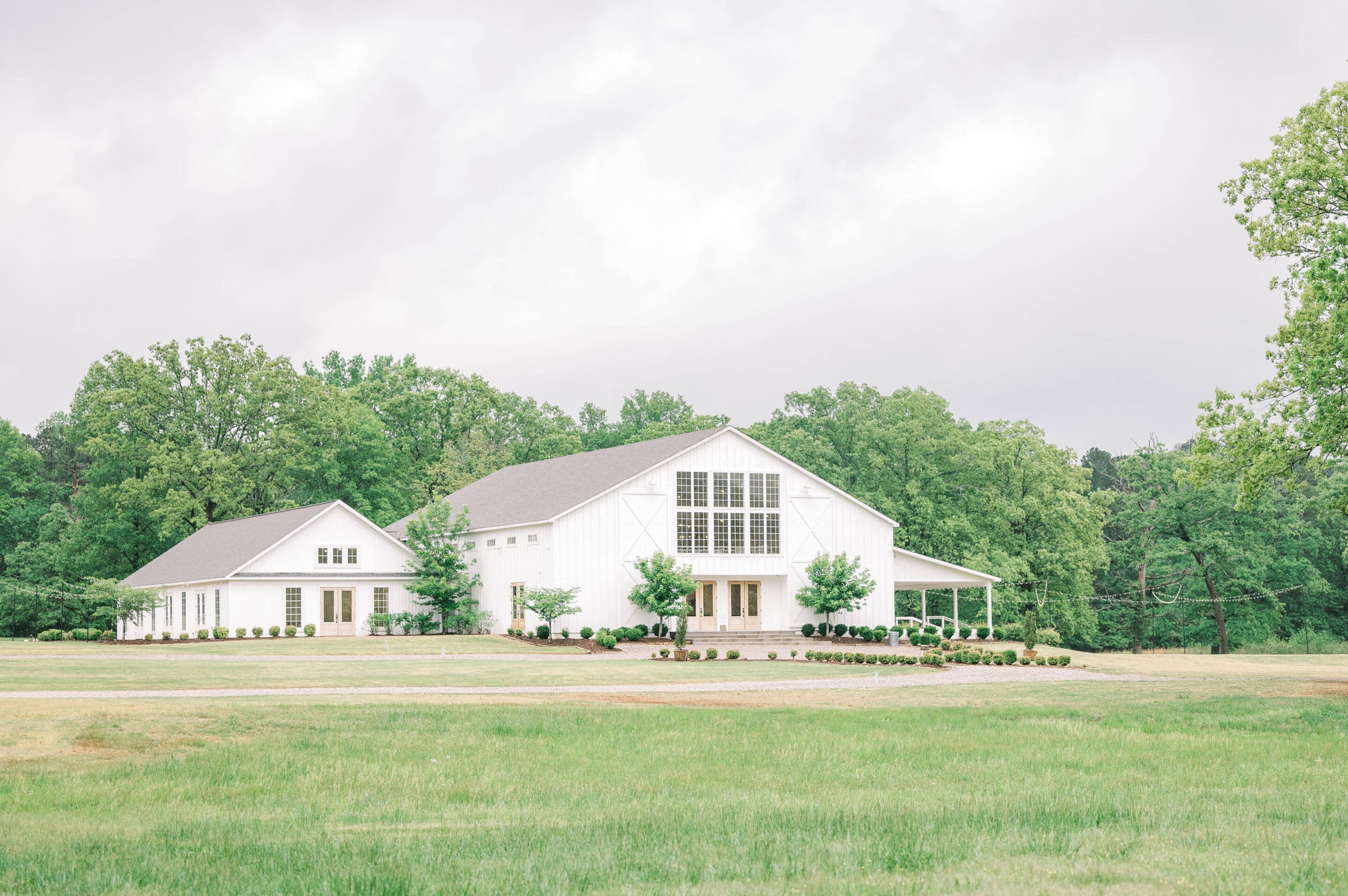 A large, modern white barn with many windows sits on a grassy lawn, surrounded by trimmed bushes and tall green trees under a cloudy sky.