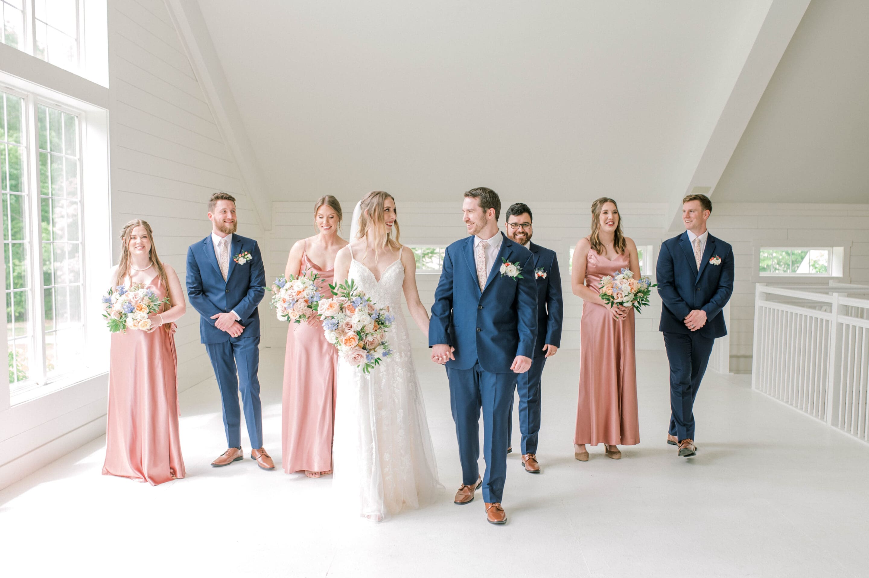 A bride and groom walk hand in hand indoors, smiling, surrounded by three bridesmaids in pink dresses and three groomsmen in navy suits, all holding pastel bouquets. The setting is bright with white walls and large windows.