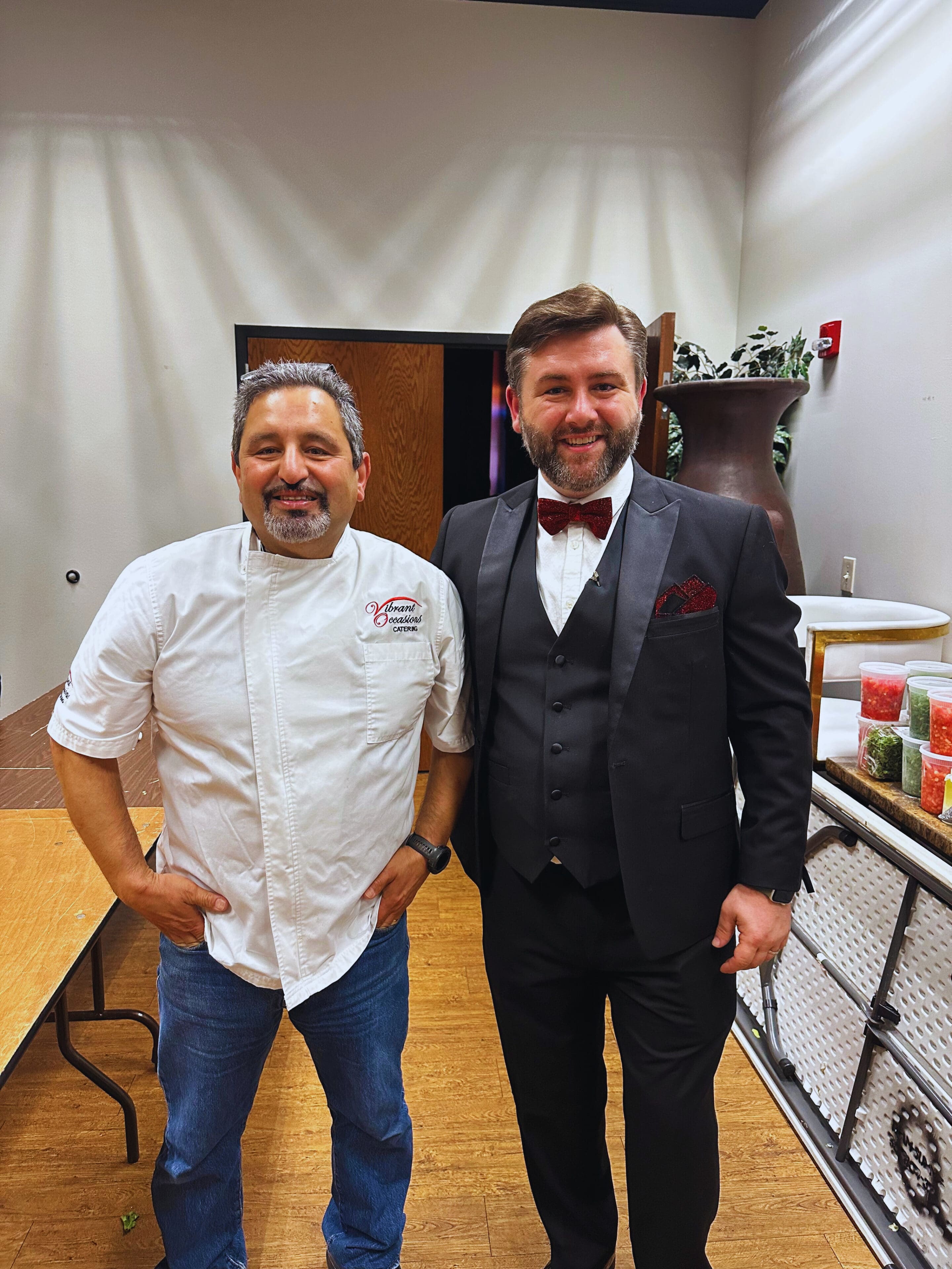 Two men stand side by side indoors. One wears a chefs jacket and jeans, while the other is dressed in a black tuxedo with a red bow tie and pocket square. They are smiling at the camera.