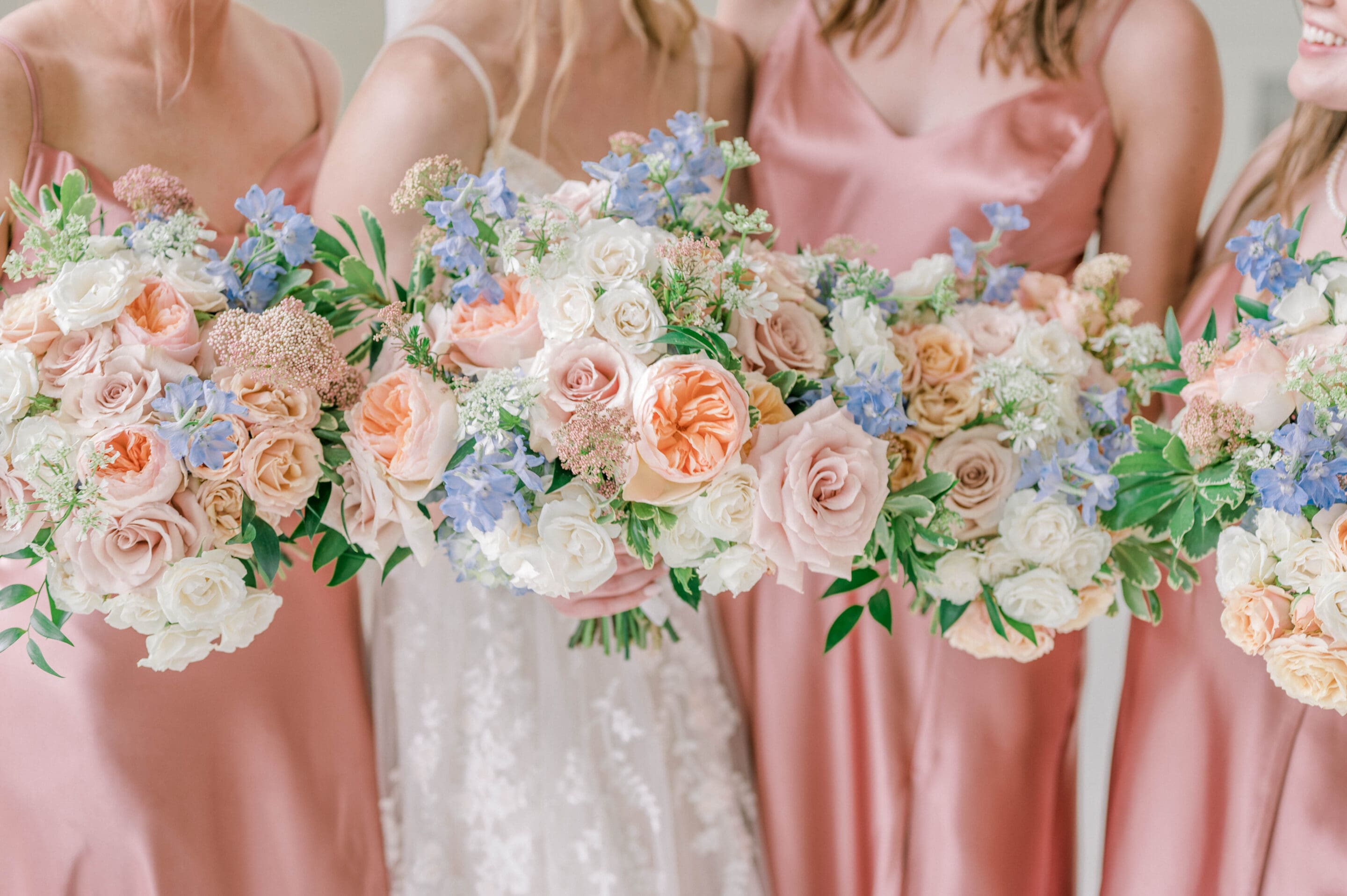 Four women in blush pink dresses hold large bouquets of pastel roses, greenery, and light blue flowers. Only their torsos are visible, and they stand closely together, smiling.