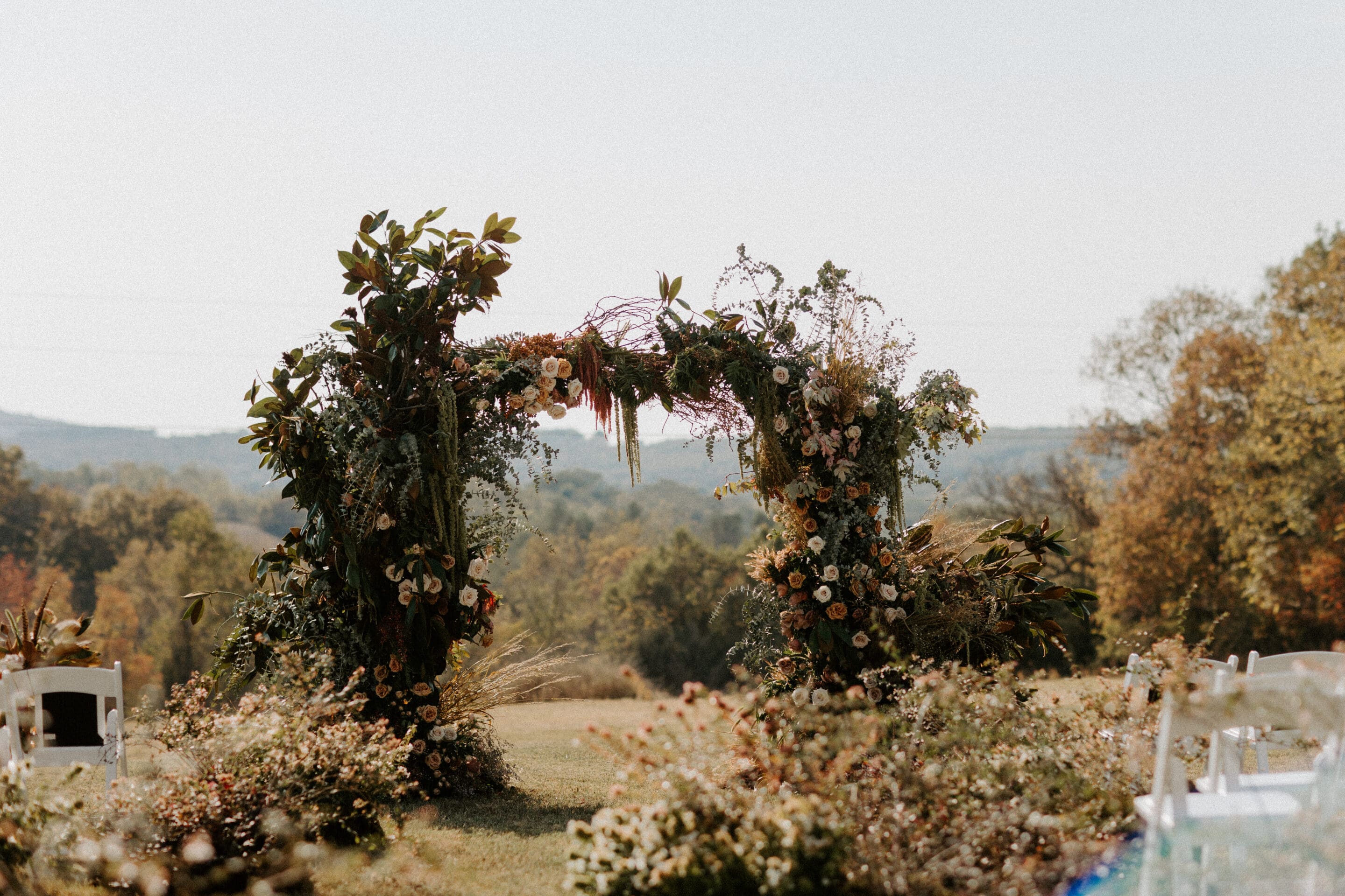 A floral wedding arch decorated with greenery and flowers stands outdoors on a grassy field, with trees and hills visible in the background under a clear sky. White chairs are set up on either side of the arch.