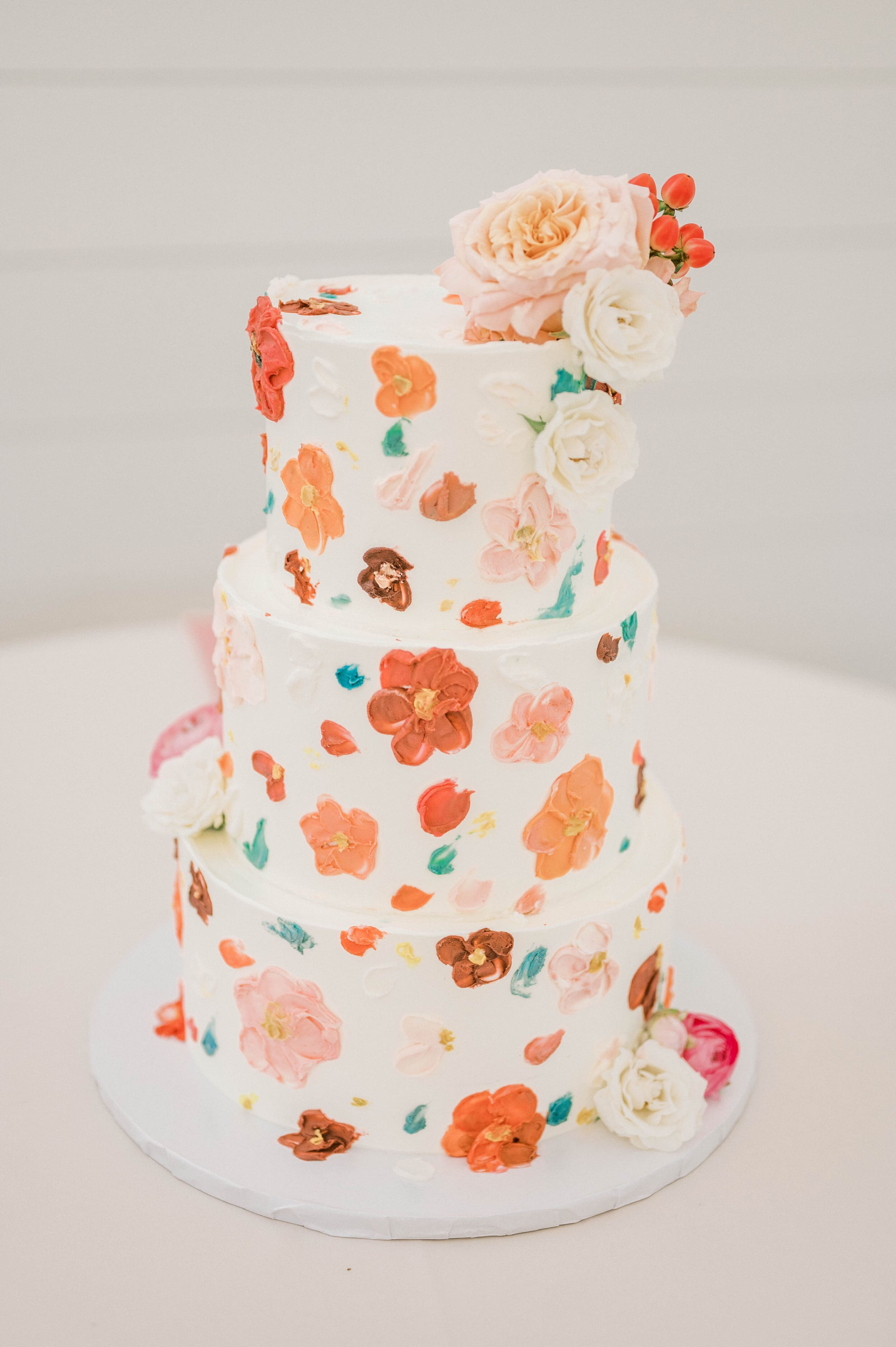 A three-tiered white cake decorated with colorful, hand-painted floral designs and topped with an arrangement of fresh flowers in shades of pink, orange, and cream.