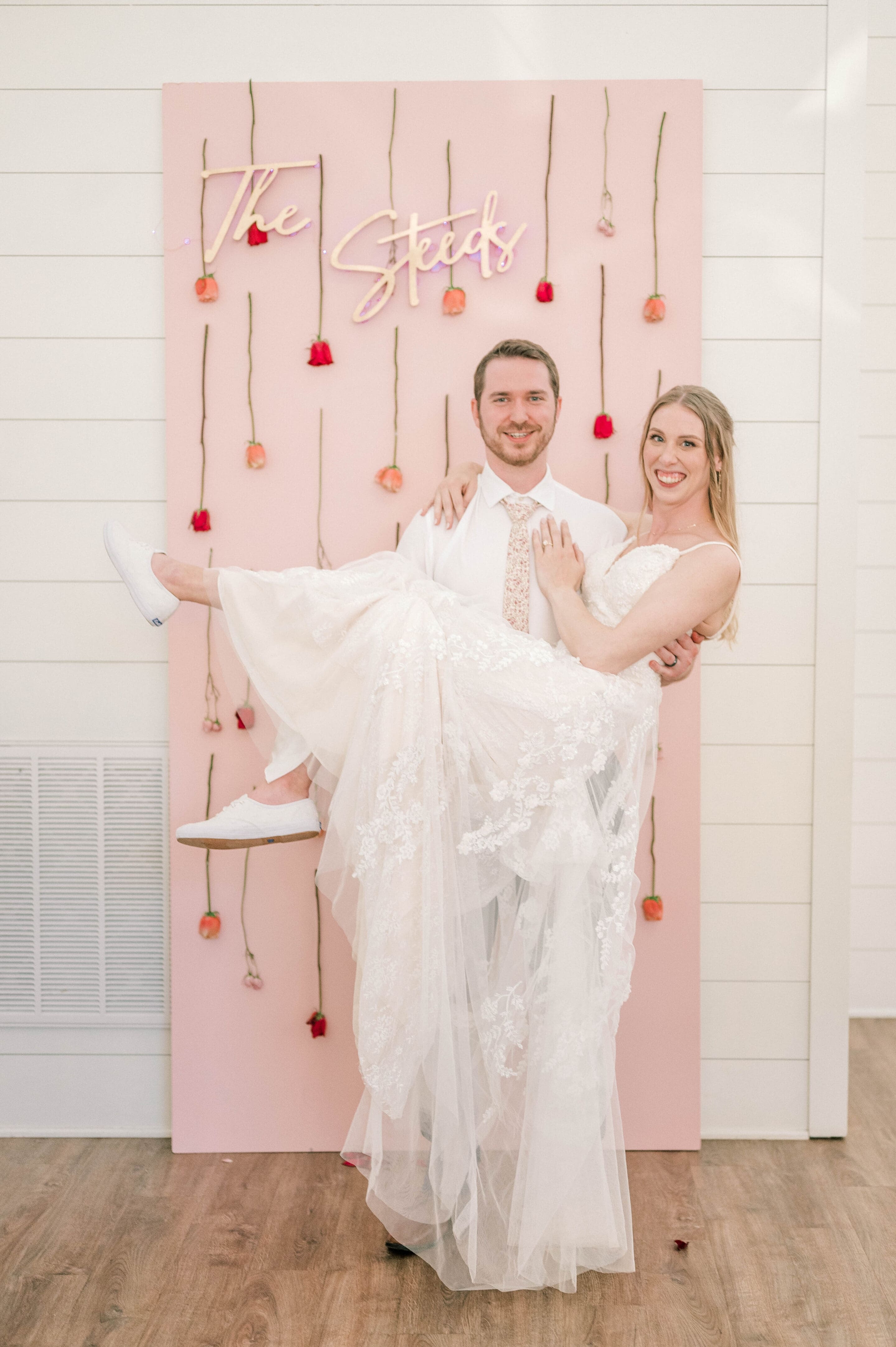 A groom in a white shirt and tie holds his smiling bride, dressed in a white gown and sneakers, in front of a pink backdrop decorated with hanging flowers and a neon sign that reads “The Steids.”.