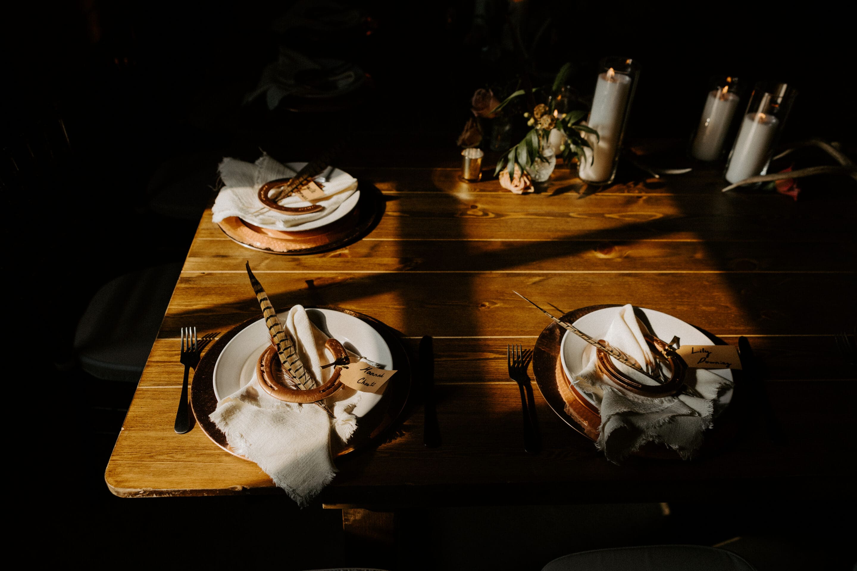 A wooden table set for three with rustic plates, napkins, and feathers as decoration. Sunlight casts shadows across the table, which also holds candles and a small floral arrangement.