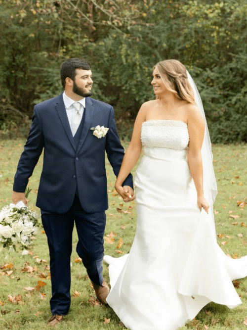 Arkansas Bride and Groom Portrait A bride and groom walk hand in hand outdoors on a grassy area. The groom wears a navy suit and holds a bouquet; the bride wears a strapless white gown and veil. They smile at each other with trees in the background.