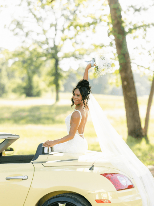 Arkansas Bride on Beautiful Car A bride in a white dress sits on the back of a yellow convertible, smiling and holding her bouquet in the air. Her veil flows behind her, and sunlight filters through the trees in the background.